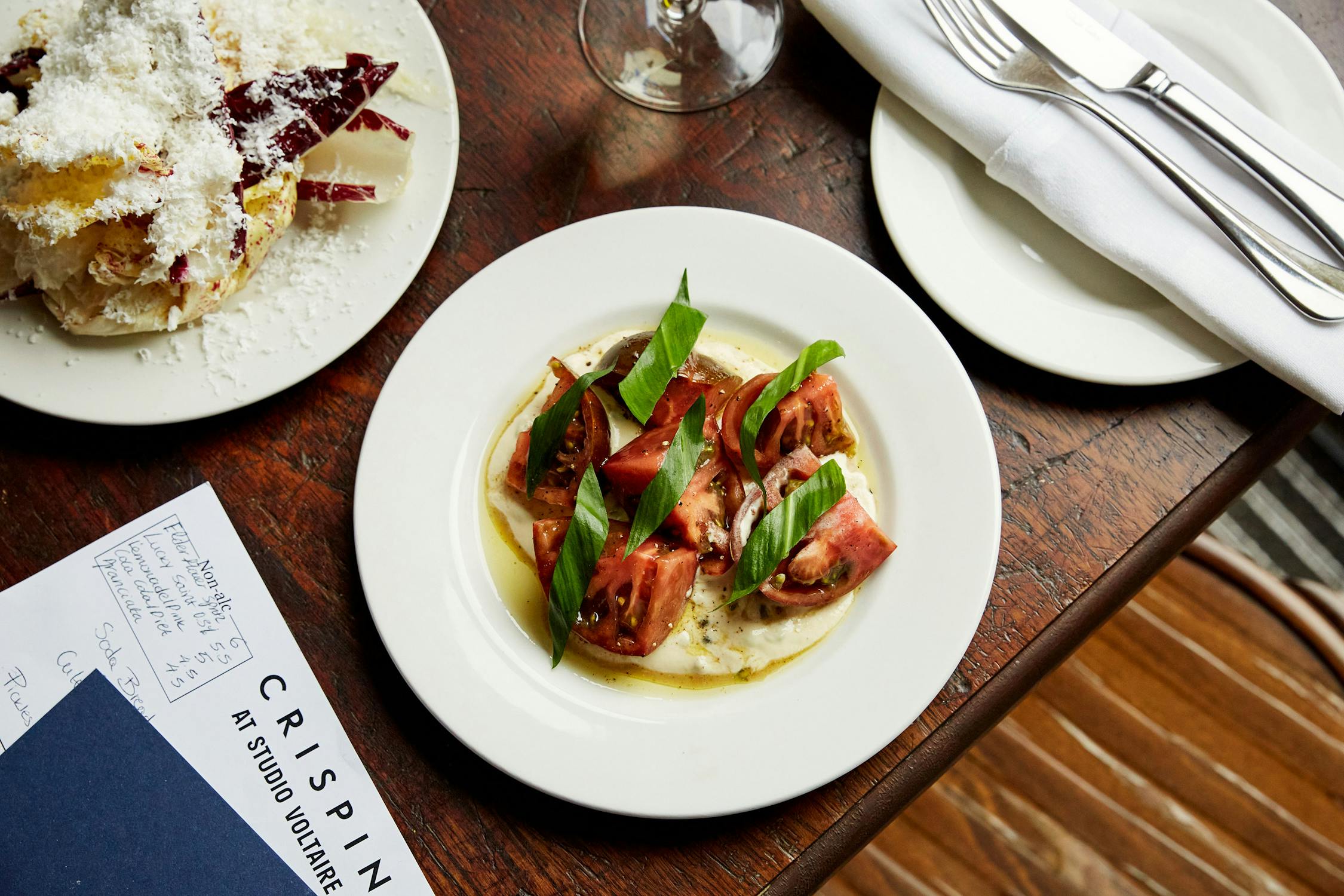 A plate of sliced tomatoes on creamy cheese, garnished with green leaves and olive oil, sits on a wooden table beside cutlery, a wine glass, a plate of dessert, and a Crispin restaurant menu.