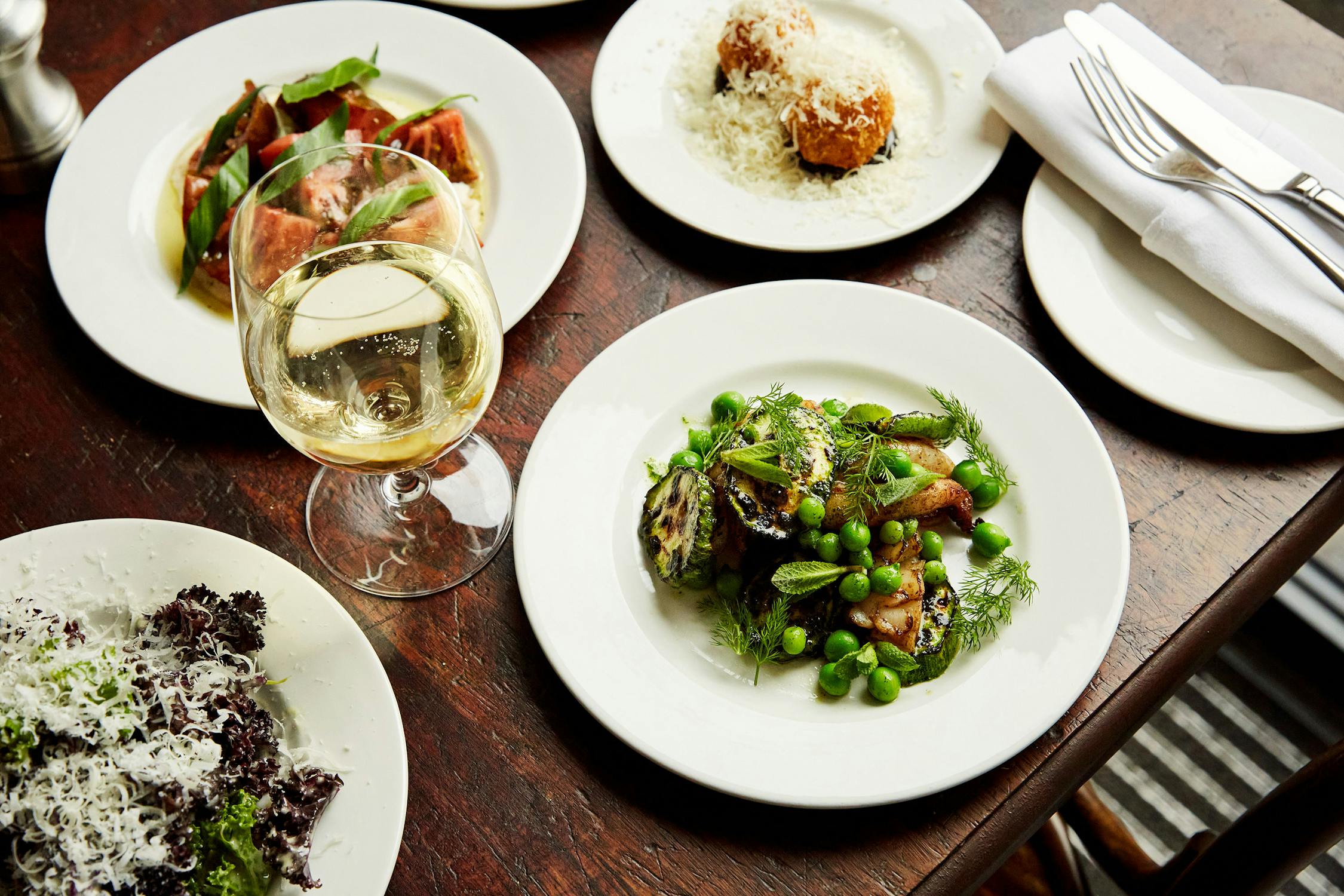 A wooden table set with plates of gourmet salads and appetizers, including a plate of green peas and vegetables, a plate of greens with grated cheese, and a glass of white wine. A napkin and utensils are on a plate nearby.