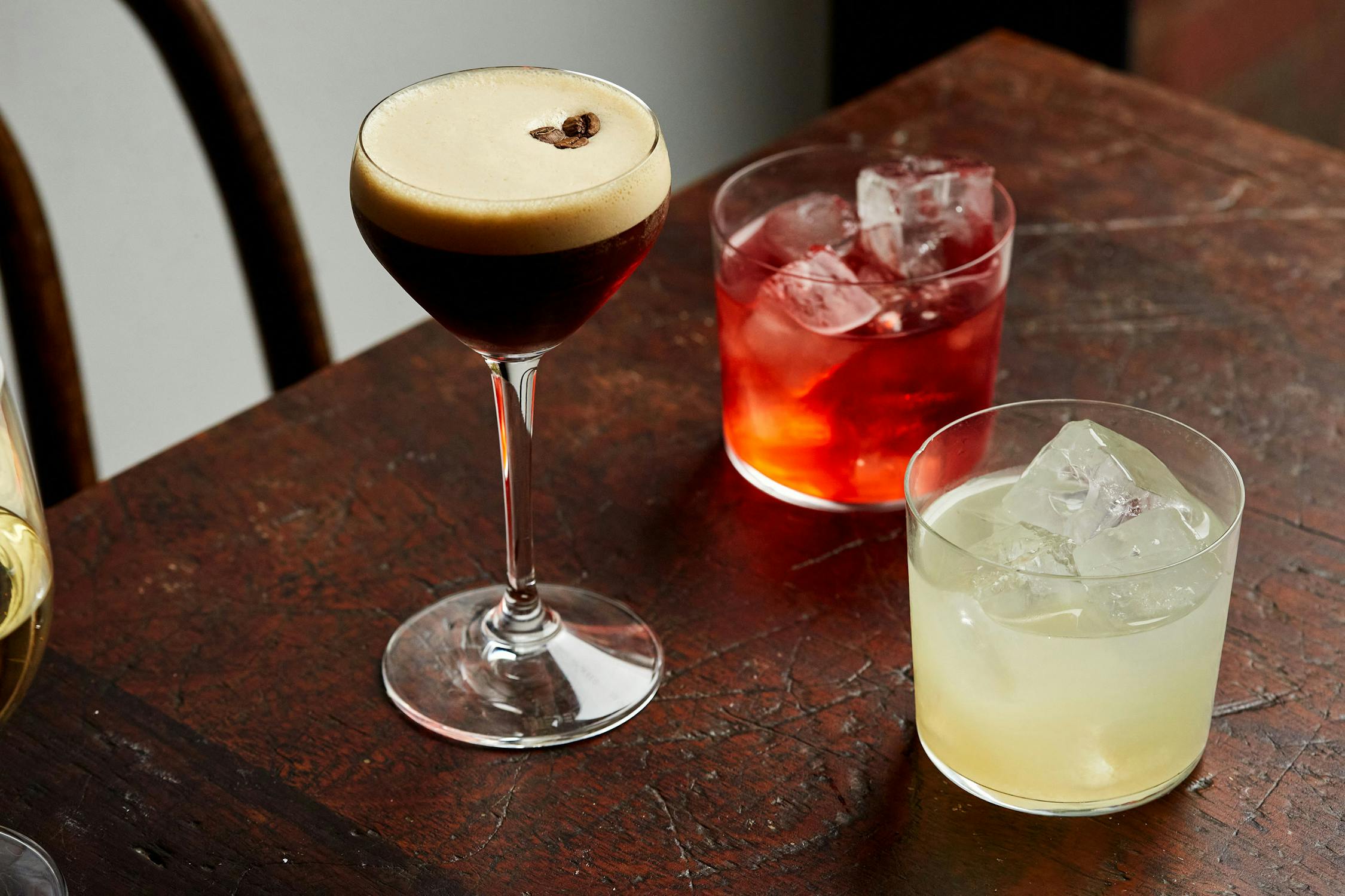 Three cocktails on a wooden table: an espresso martini with foam and coffee bean garnish, a red cocktail on ice, and a pale yellow cocktail on ice. Part of a fourth drink is partially visible on the left edge.