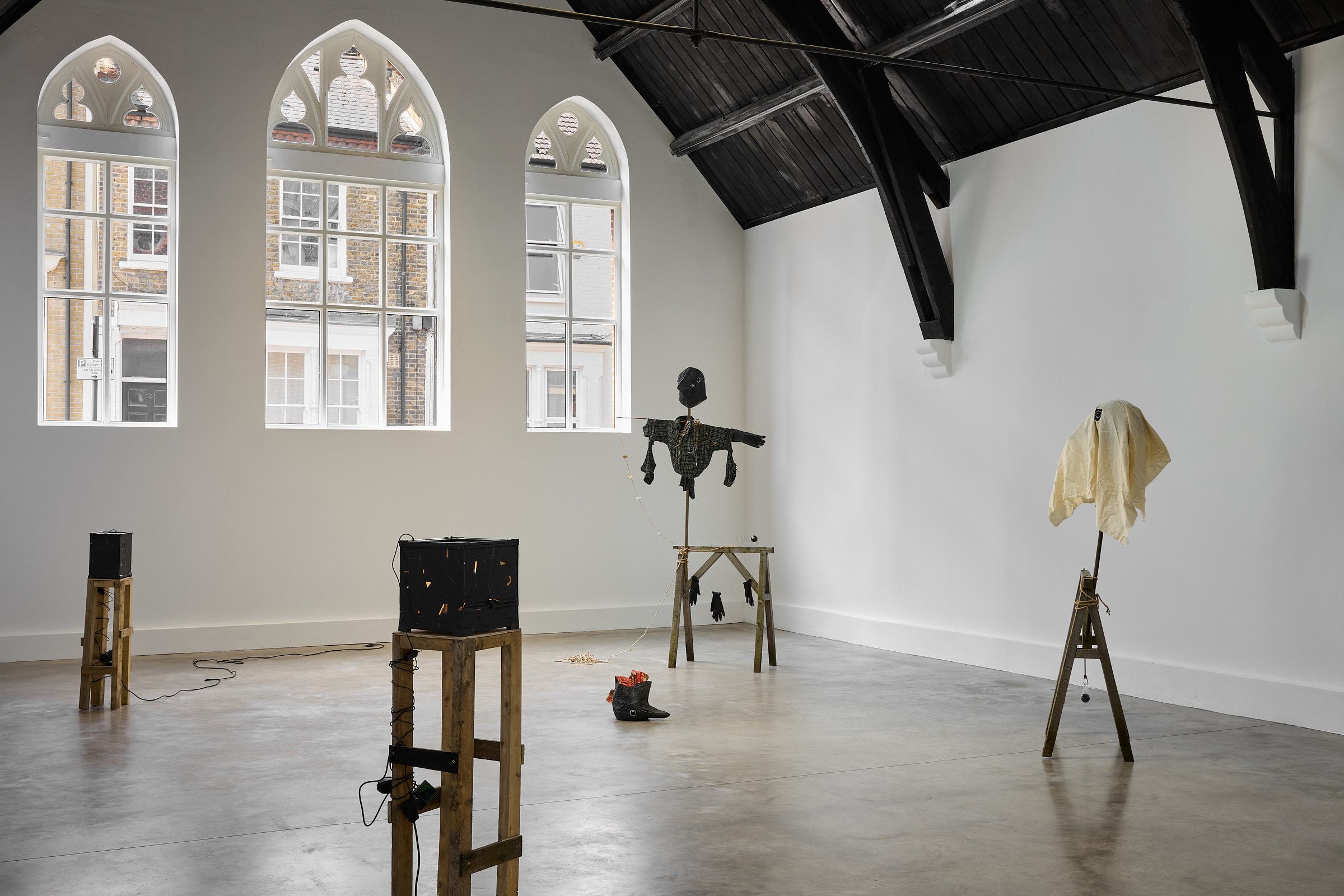 A contemporary art gallery with arched windows and black wooden beams features sculptures by Caspar Heinemann on wooden stands, including a black scarecrow and a draped figure, in a spacious, minimalist setting.