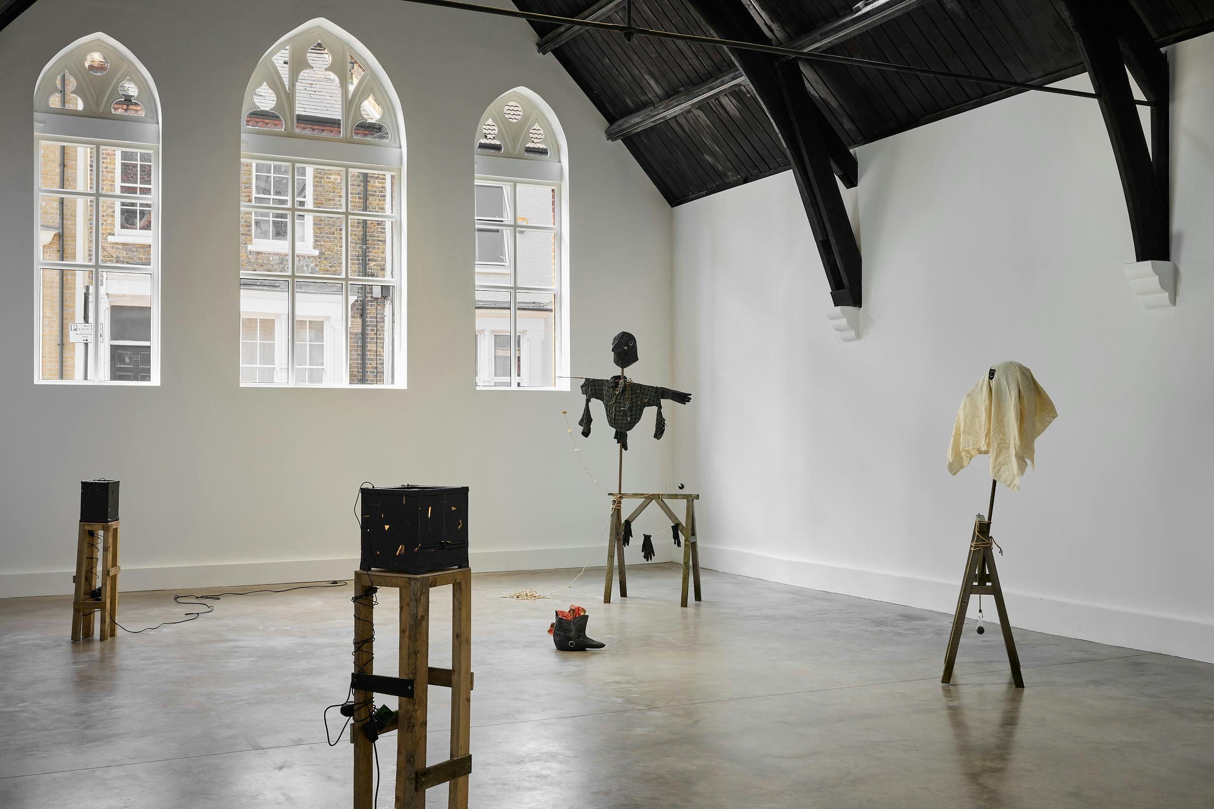 A contemporary art gallery with arched windows and black wooden beams features sculptures by Caspar Heinemann on wooden stands, including a black scarecrow and a draped figure, in a spacious, minimalist setting.