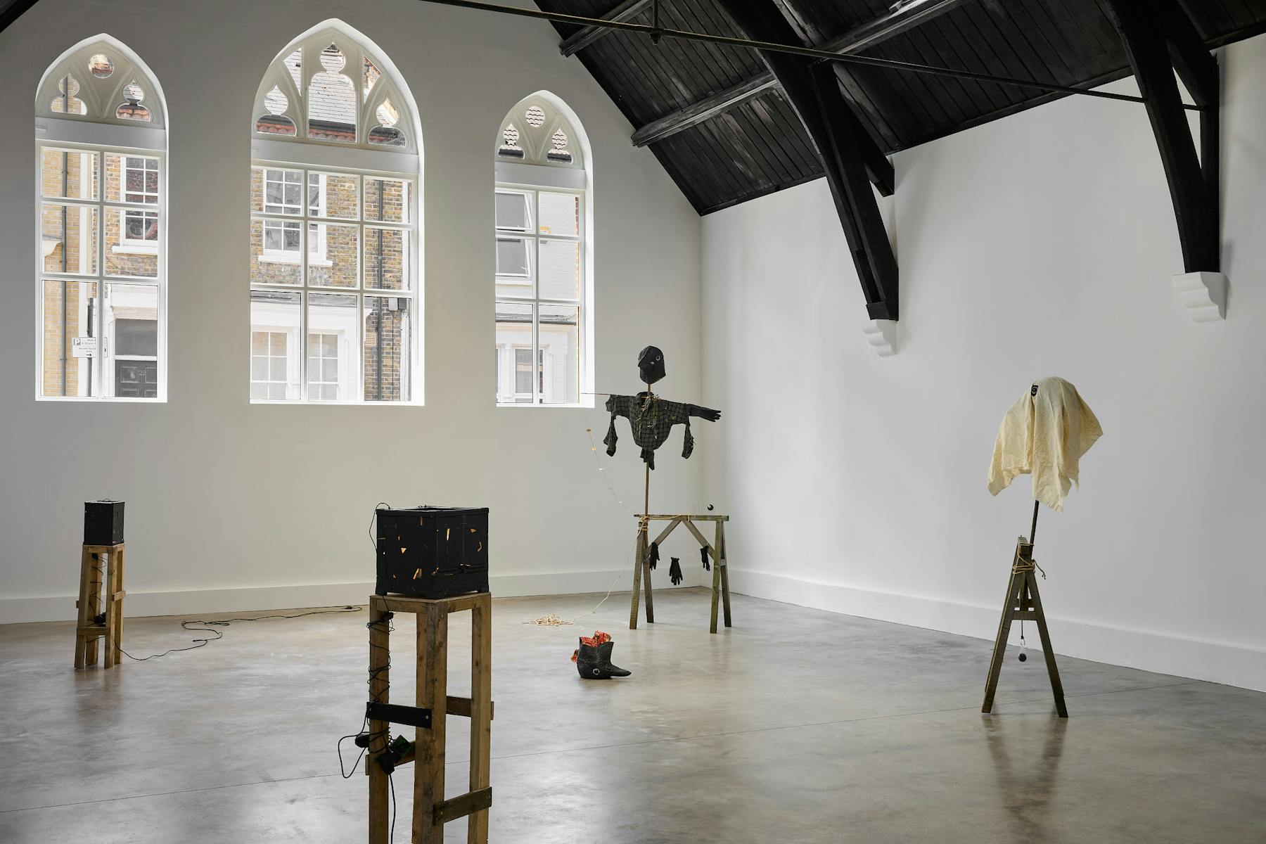 A contemporary art gallery with arched windows and black wooden beams features sculptures by Caspar Heinemann on wooden stands, including a black scarecrow and a draped figure, in a spacious, minimalist setting.