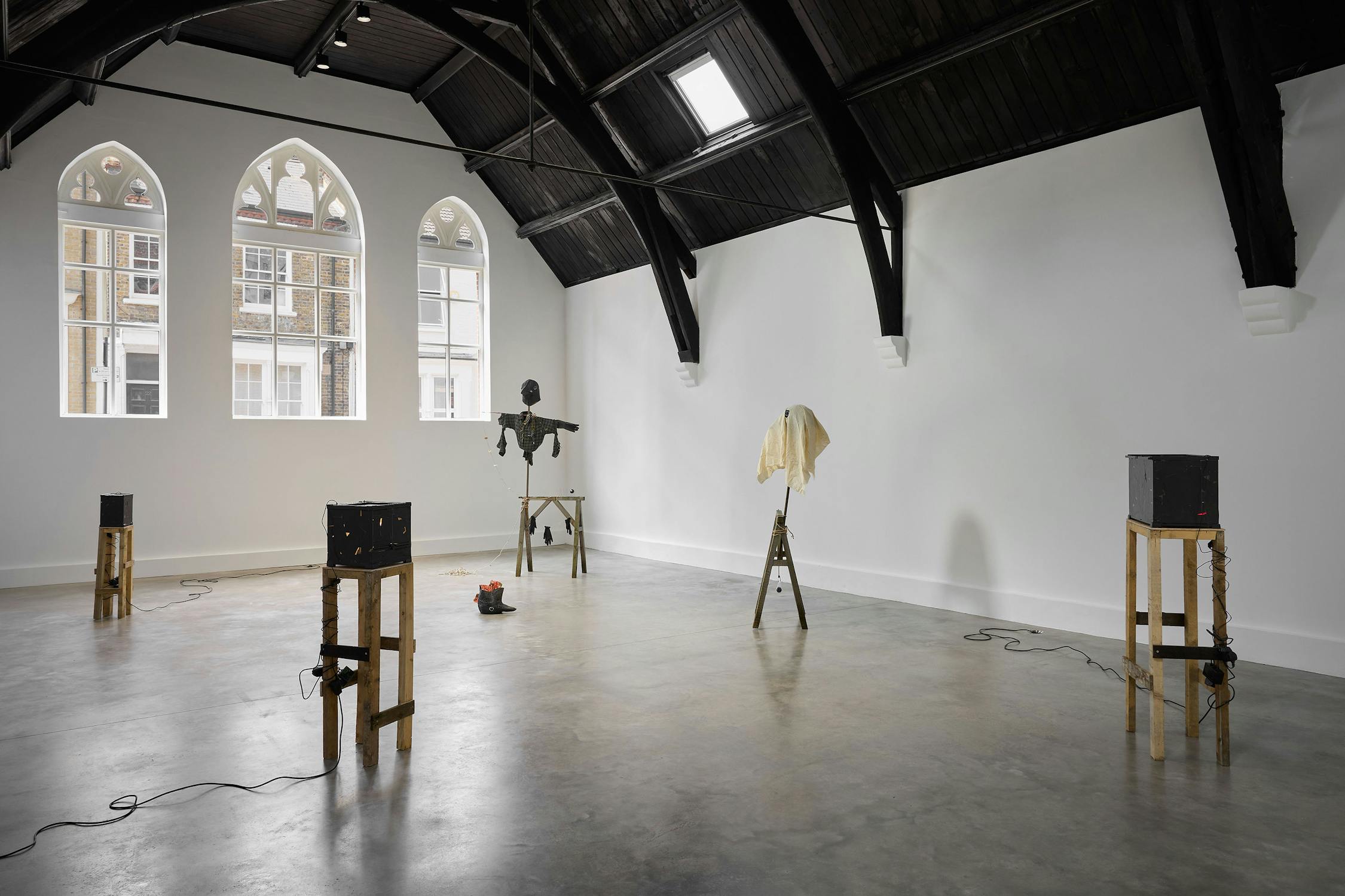 A gallery with high dark-beamed ceilings and arched windows features four wooden stands with abstract sculptures and electronics on them, placed around a spacious, light-filled concrete floor.