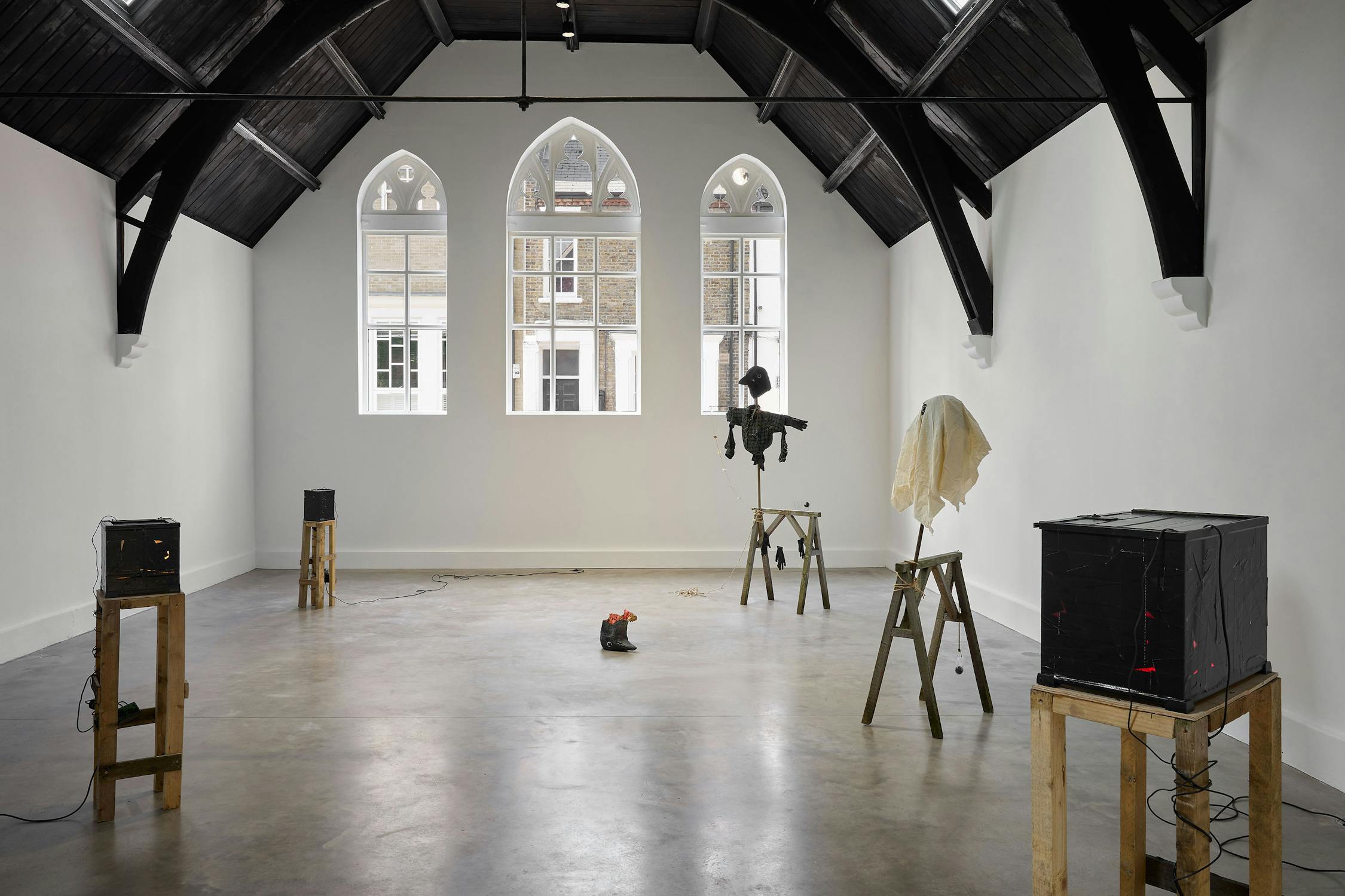 Art installation in a spacious, white-walled room with arched windows and a high, dark-beamed ceiling. Four mixed-media sculptures sit on wooden stands, with one object on the floor in the centre. Natural light fills the space.