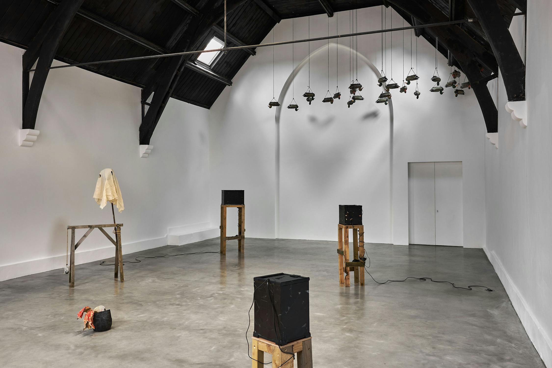 A minimalist art installation in a white gallery with high, black-beamed ceilings; features wooden stands, black boxes with cables, a draped white cloth, small hanging sculptures, and a black and red mask sculpture on the floor.