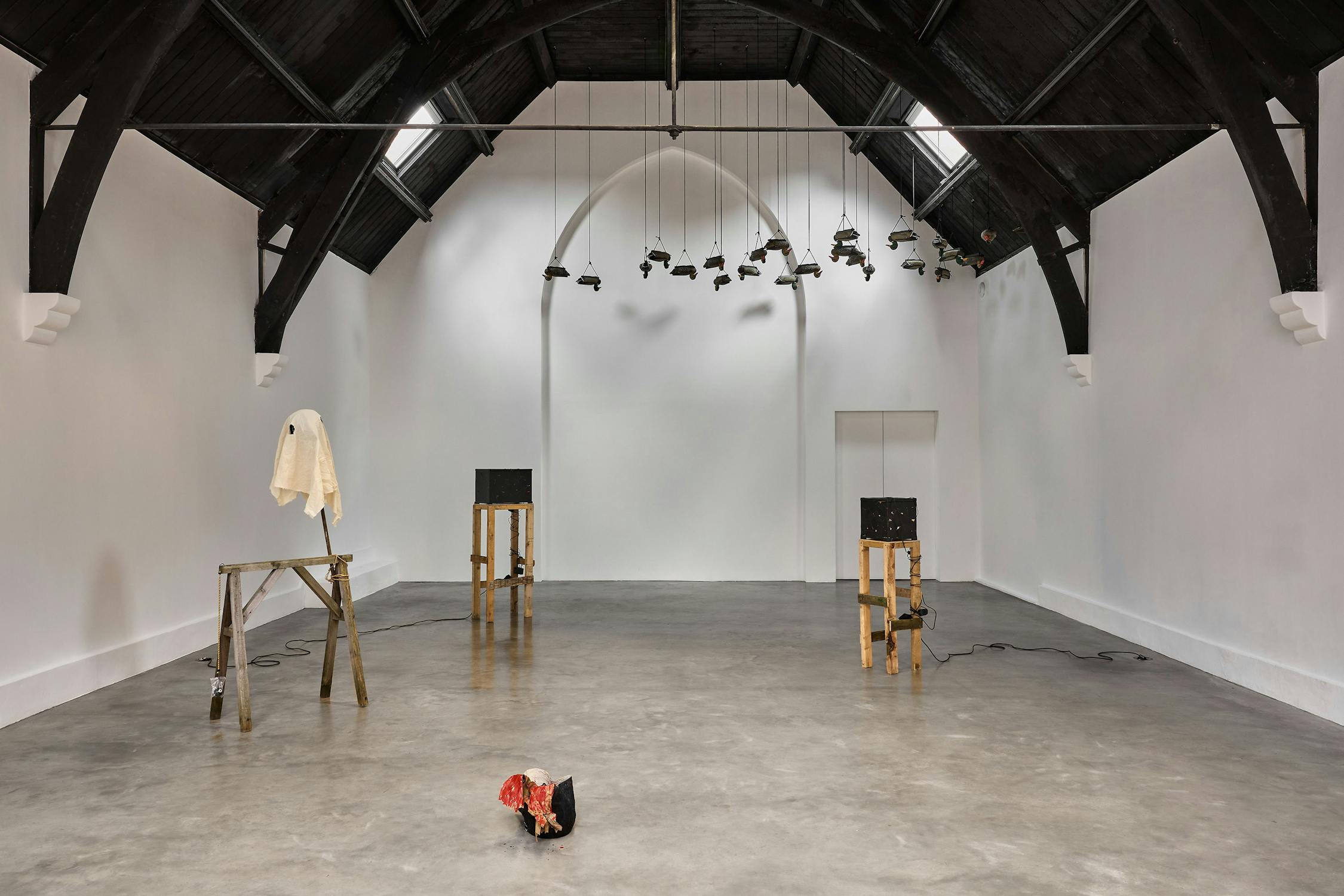 An art gallery with a high, dark vaulted ceiling features minimalistic sculptures: a ghost-like figure on a sawhorse, a small black bird mask stuffed with red paper on the floor, and plastic ducks suspended from the ceiling.