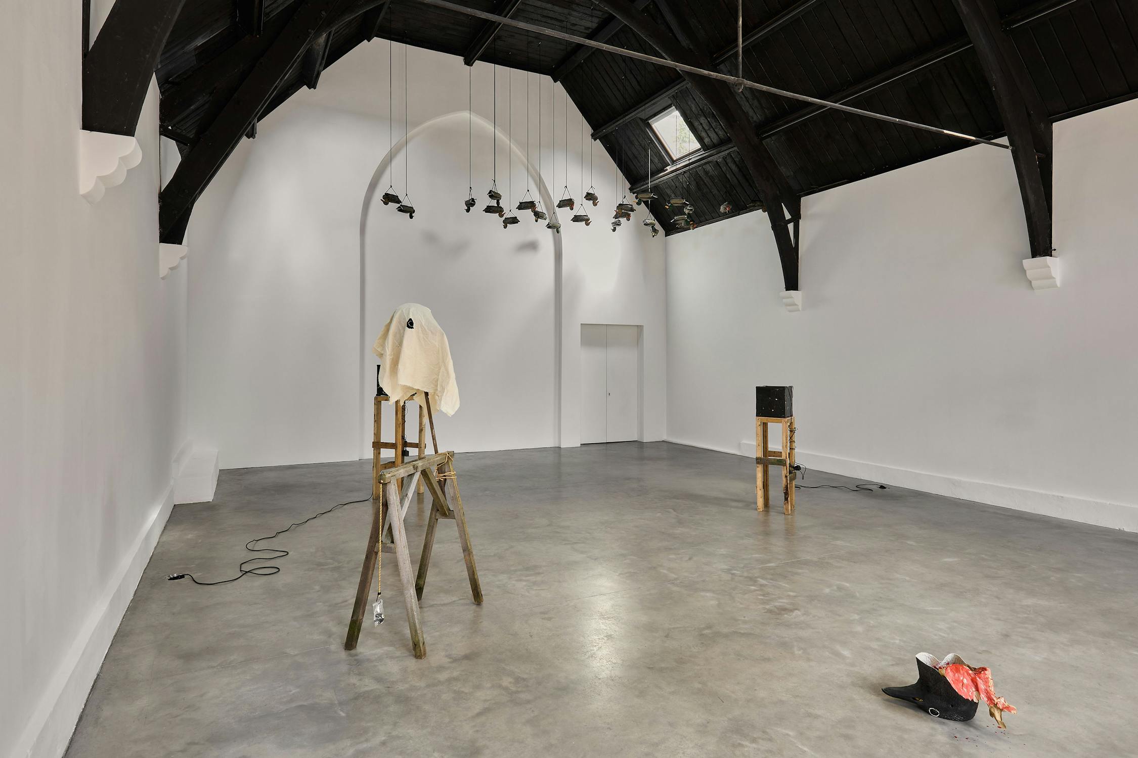 Art installation in a spacious, white-walled room with a high, dark arched ceiling. A wooden structure draped with a white cloth stands in the centre, with plastic ducks above and scattered objects on the floor.