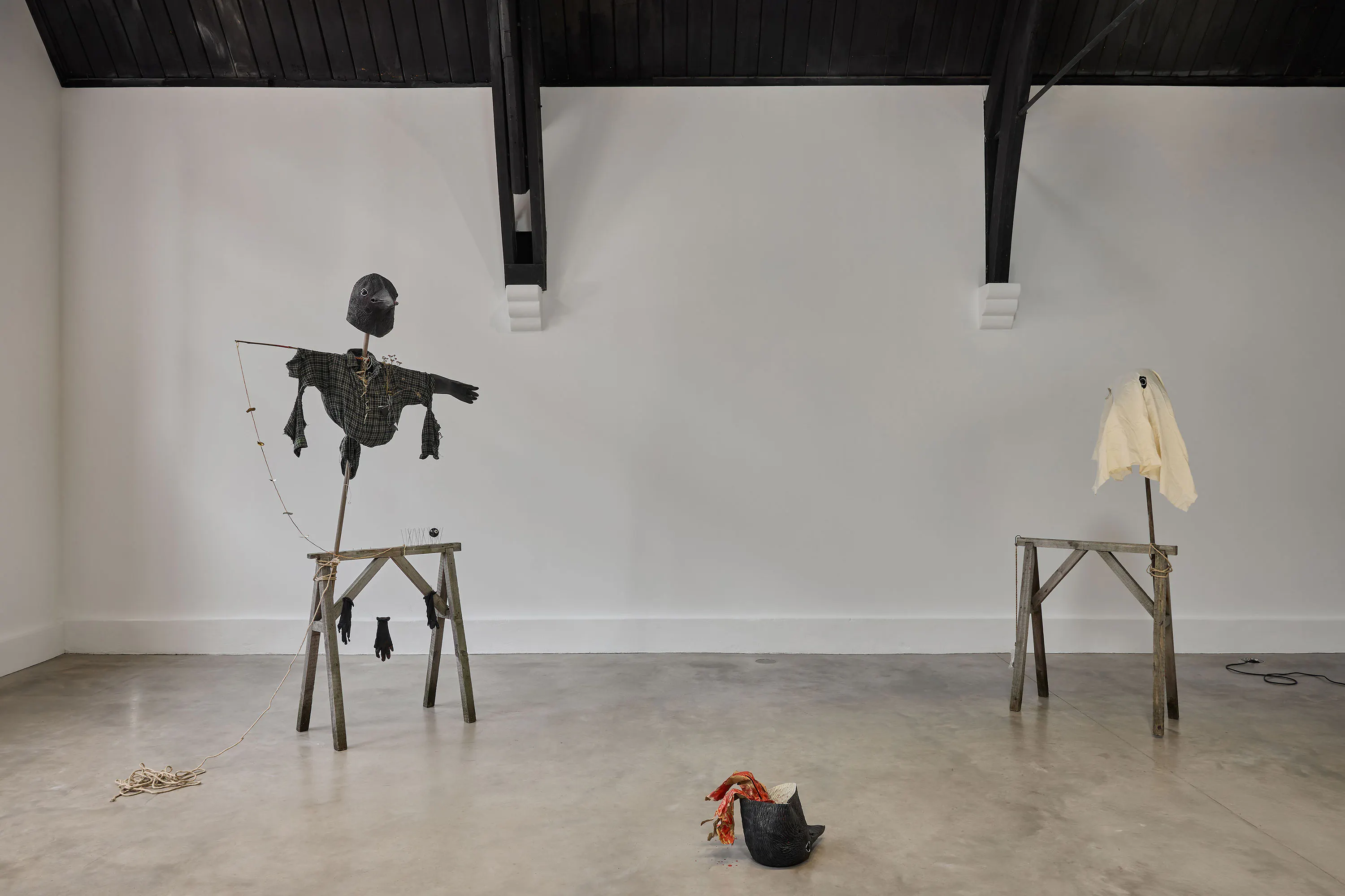 Two wooden and fabric sculptures reminiscent of makeshift scarecrows: one with dark clothes and a rubber crow mask, the other with a crow mask draped in white fabric. Objects, including a rope, another mask, and fabric scraps, lie on the polished concrete floor between them.