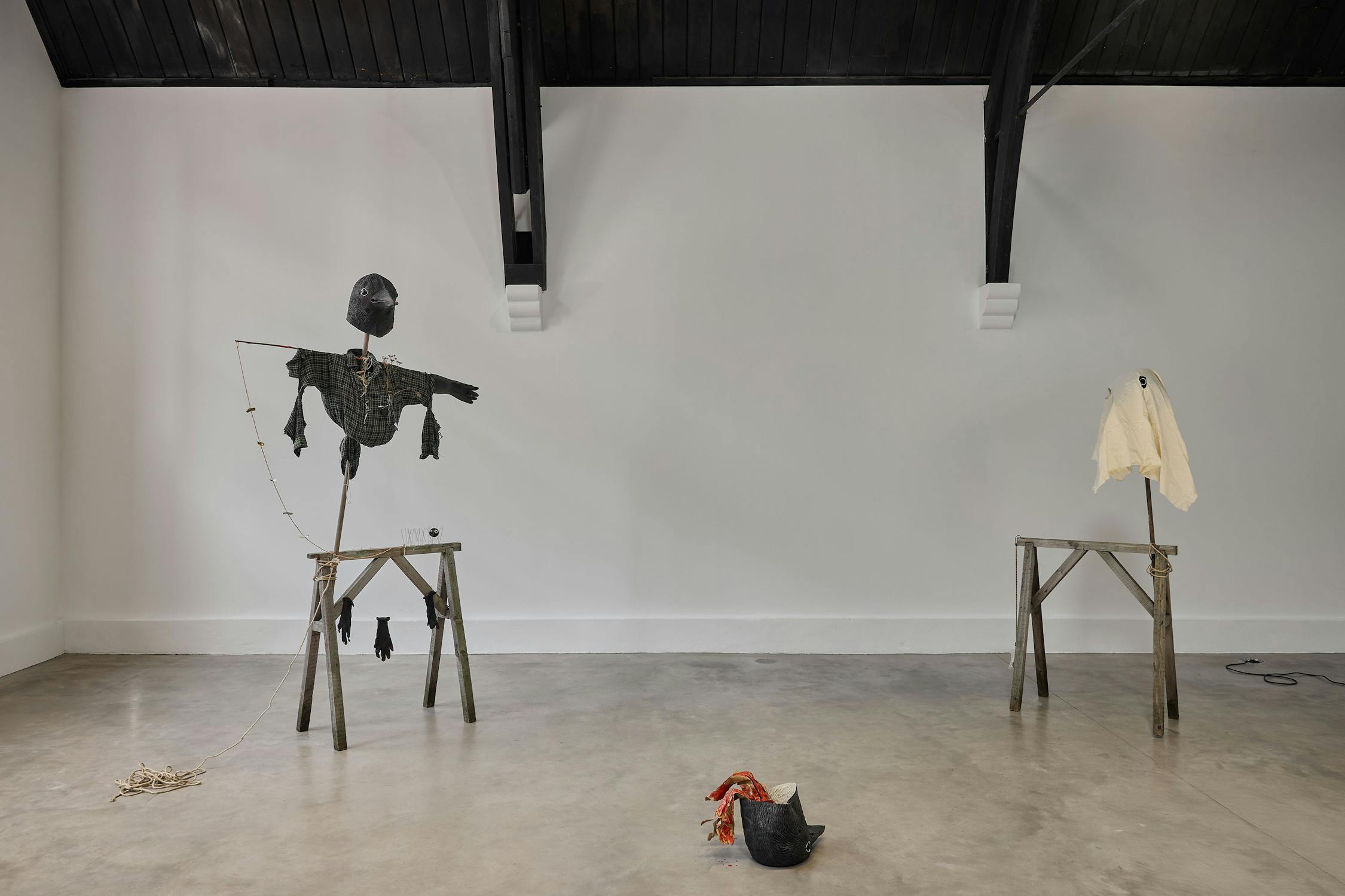 Two wooden and fabric sculptures reminiscent of makeshift scarecrows: one with dark clothes and a rubber crow mask, the other with a crow mask draped in white fabric. Objects, including a rope, another mask, and fabric scraps, lie on the polished concrete floor between them.