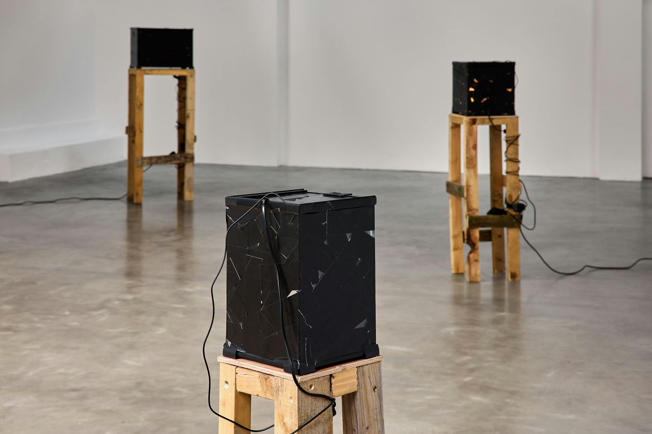 Three black cube-shaped objects with wires sit on tall wooden stands in a minimalist gallery with a polished concrete floor and white walls.