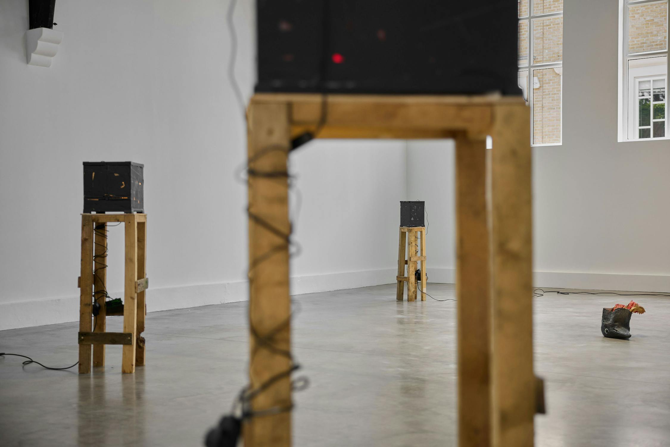 Three wooden stands with black boxes on top are spaced apart in a minimal, bright gallery room with white walls, large windows, and a concrete floor. A cord and a black mask sclupture are visible on the floor.