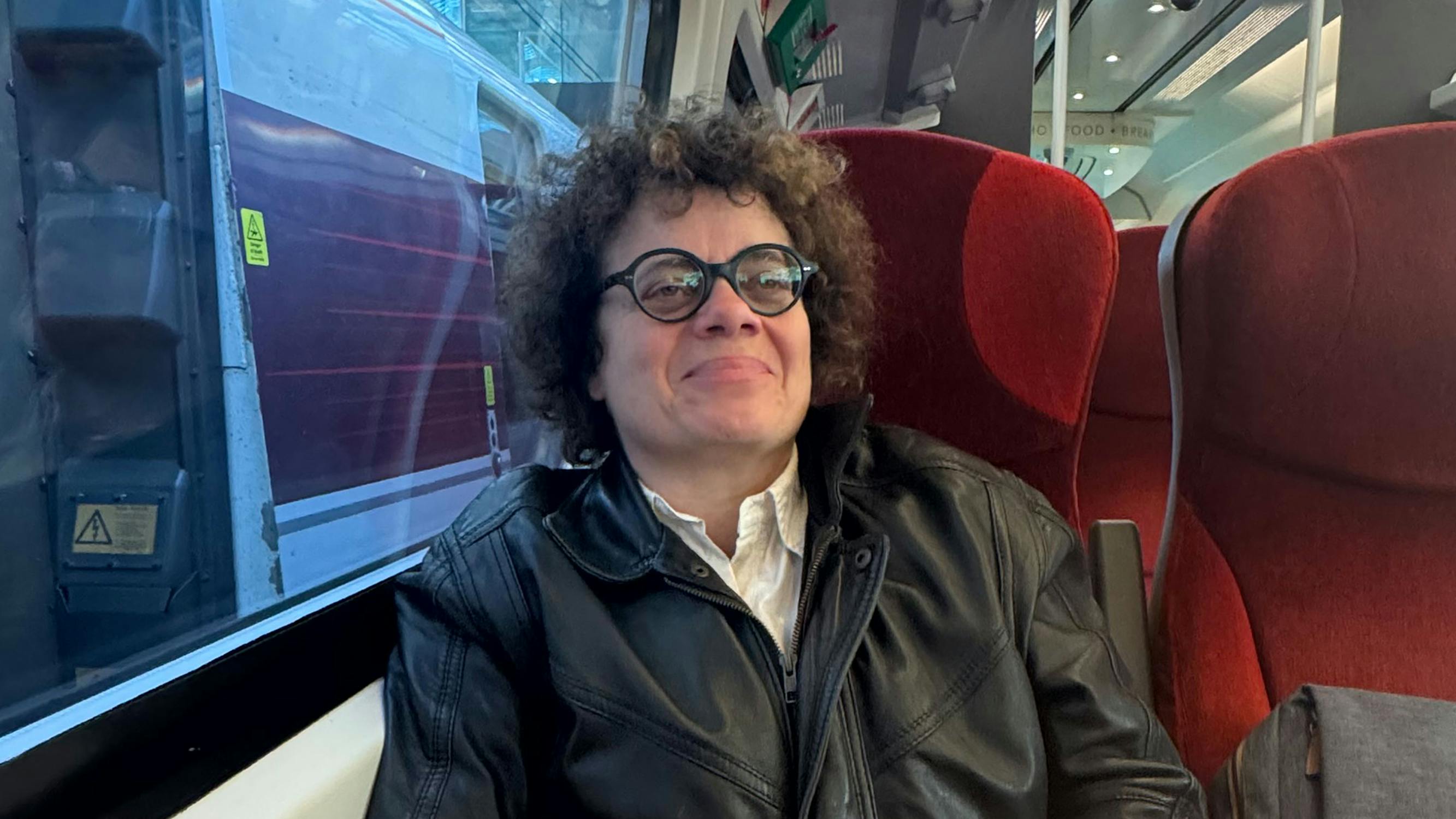 Portrait of Oreet Ashery. A person with curly hair, glasses, and a black leather jacket sits on a red train seat by the window, smiling slightly, with another train visible outside.