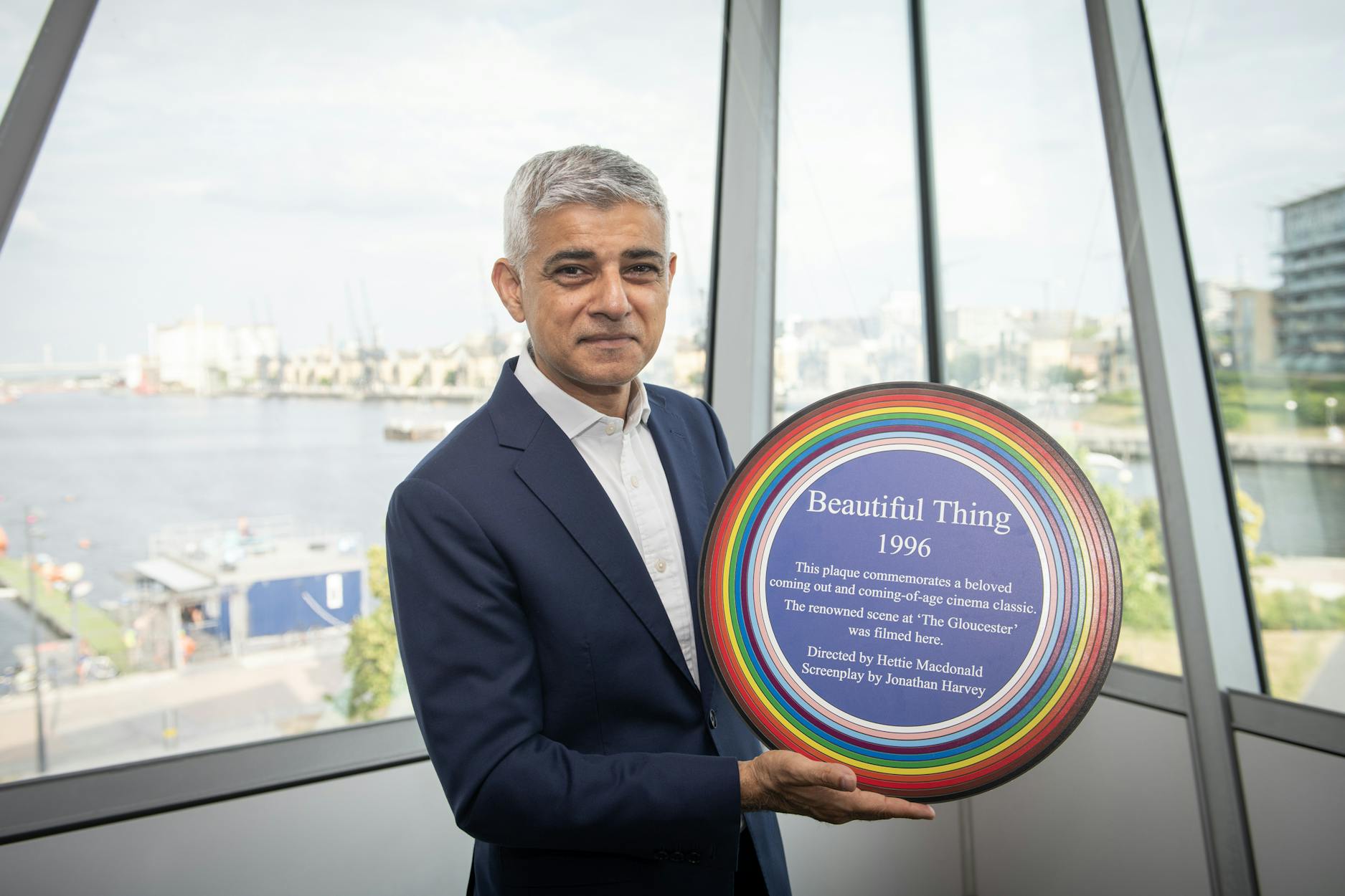 Mayor of London Sadiq Khan wearing a navy blue suit. He stands indoors in front of large glass windows overlooking a river and cityscape. He is holding a circular rainbow-coloured plaque that reads: 'Beautiful Thing, 1996.