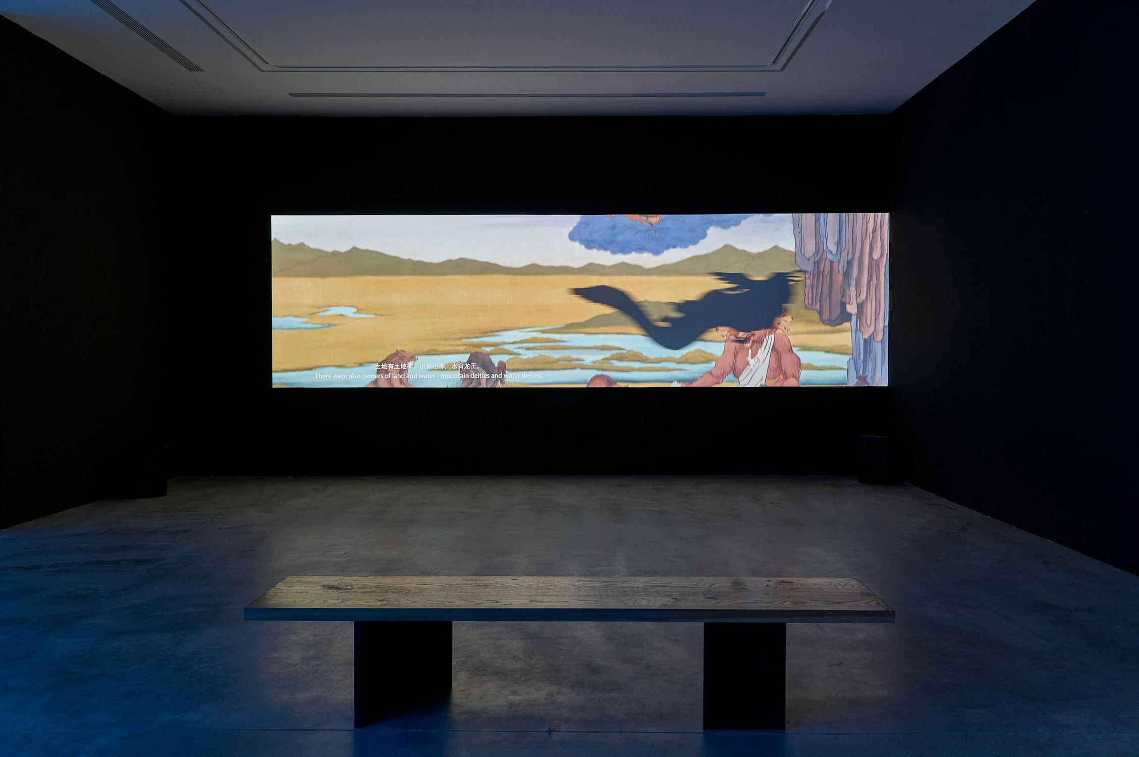 A dark gallery room features a wooden bench facing a wide projection of a surreal landscape painting, showing a desert, mountains, figures, and blue sky on the far wall.