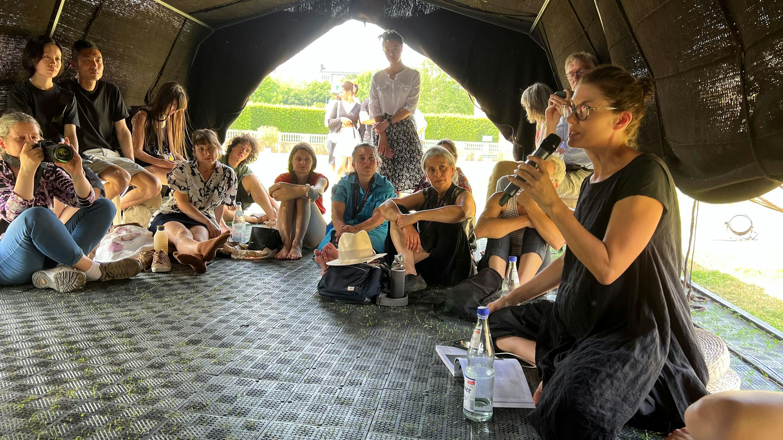 A woman speaks into a microphone while seated on the floor inside a tent, surrounded by a group of people listening attentively. Sunlight shines through the tent opening behind them.