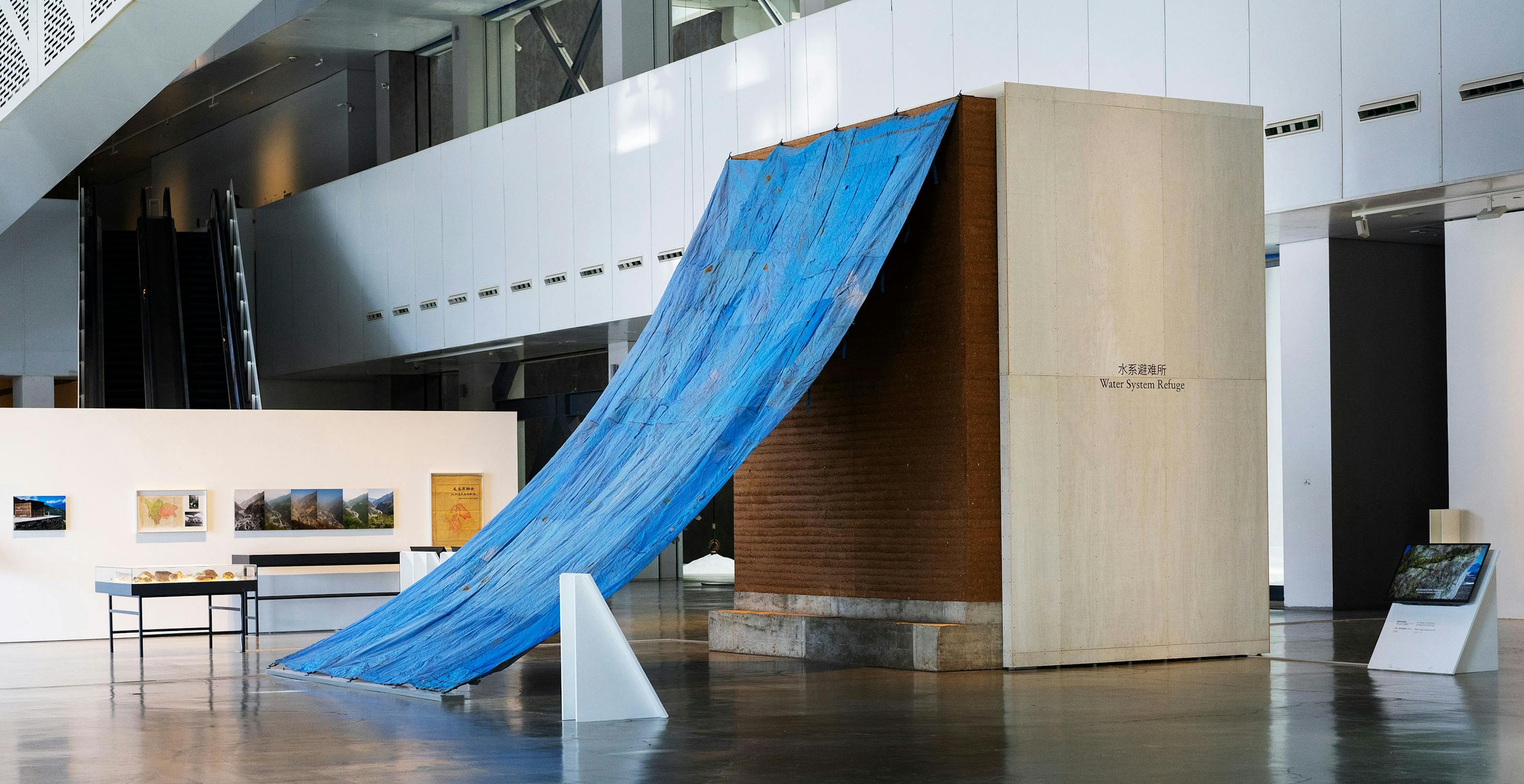 A large blue tarp cascades from the roof of a tall, rectangular structure inside a modern gallery. Framed artworks and a table are displayed nearby. The space is bright with high ceilings and contemporary design.