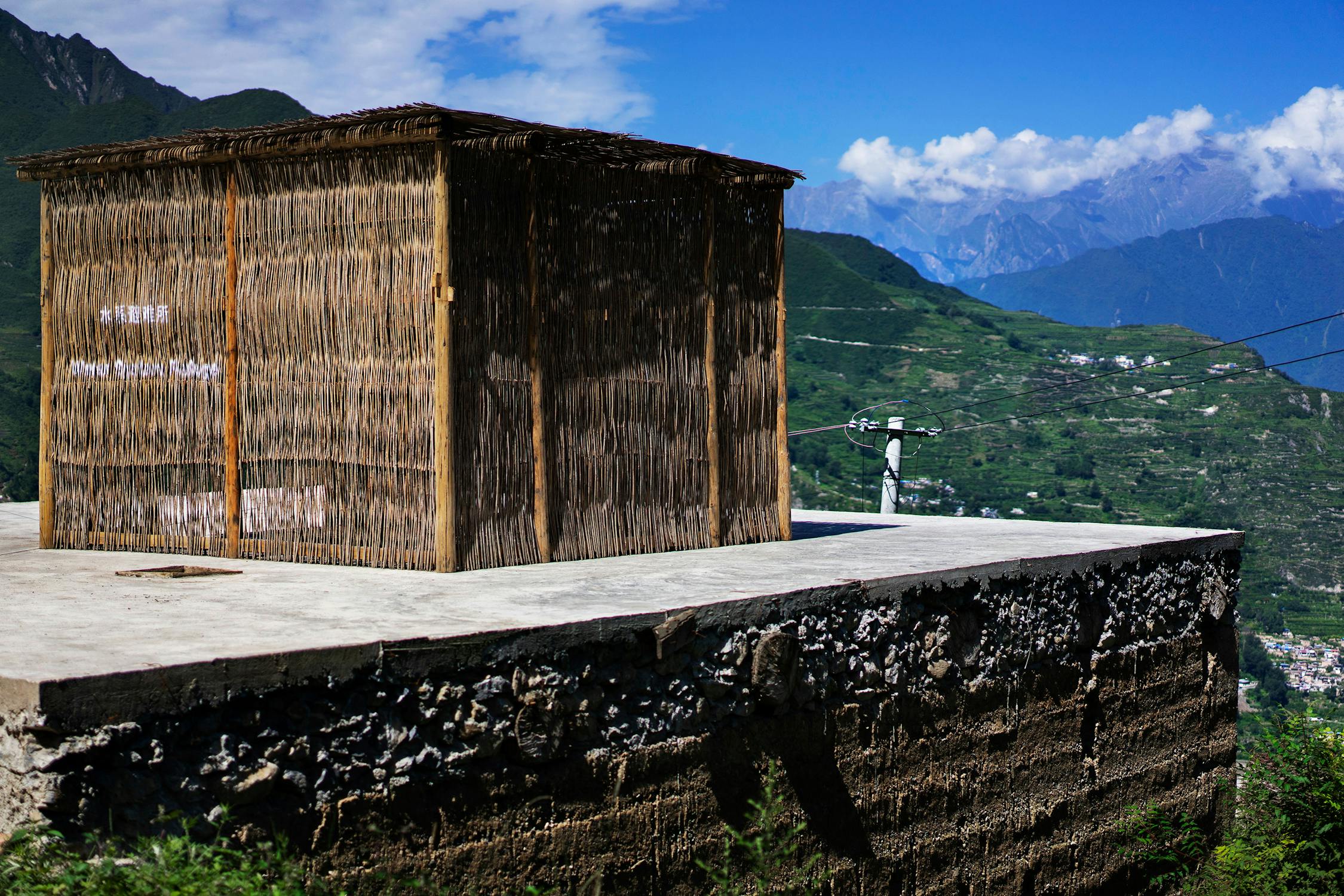 A small structure made of woven reeds stands on a concrete platform, overlooking a lush green valley with distant mountains under a blue sky.