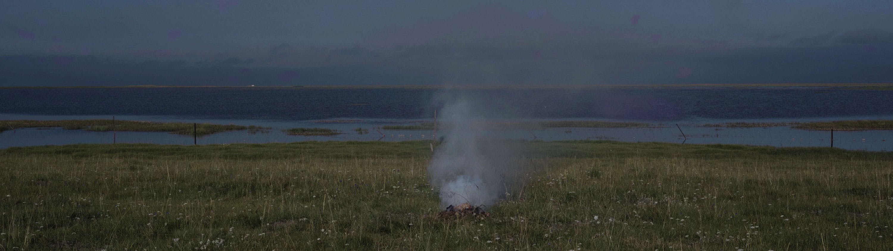 A small campfire emits smoke on a grassy field near a body of water, with a cloudy blue sky and distant shoreline in the background.