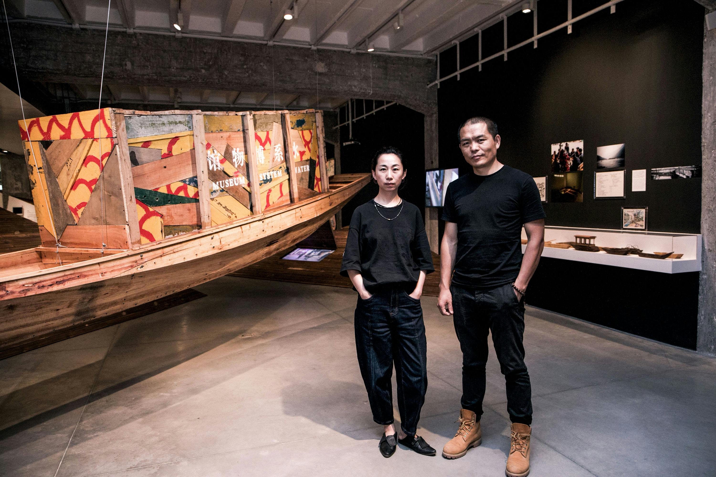Cao Minghao and Chen Jianjun stand in a modern art gallery with concrete floors, beside a large wooden boat installation decorated with colourful patterns. The walls display photographs and other artworks. Both wear black clothing and face the camera.