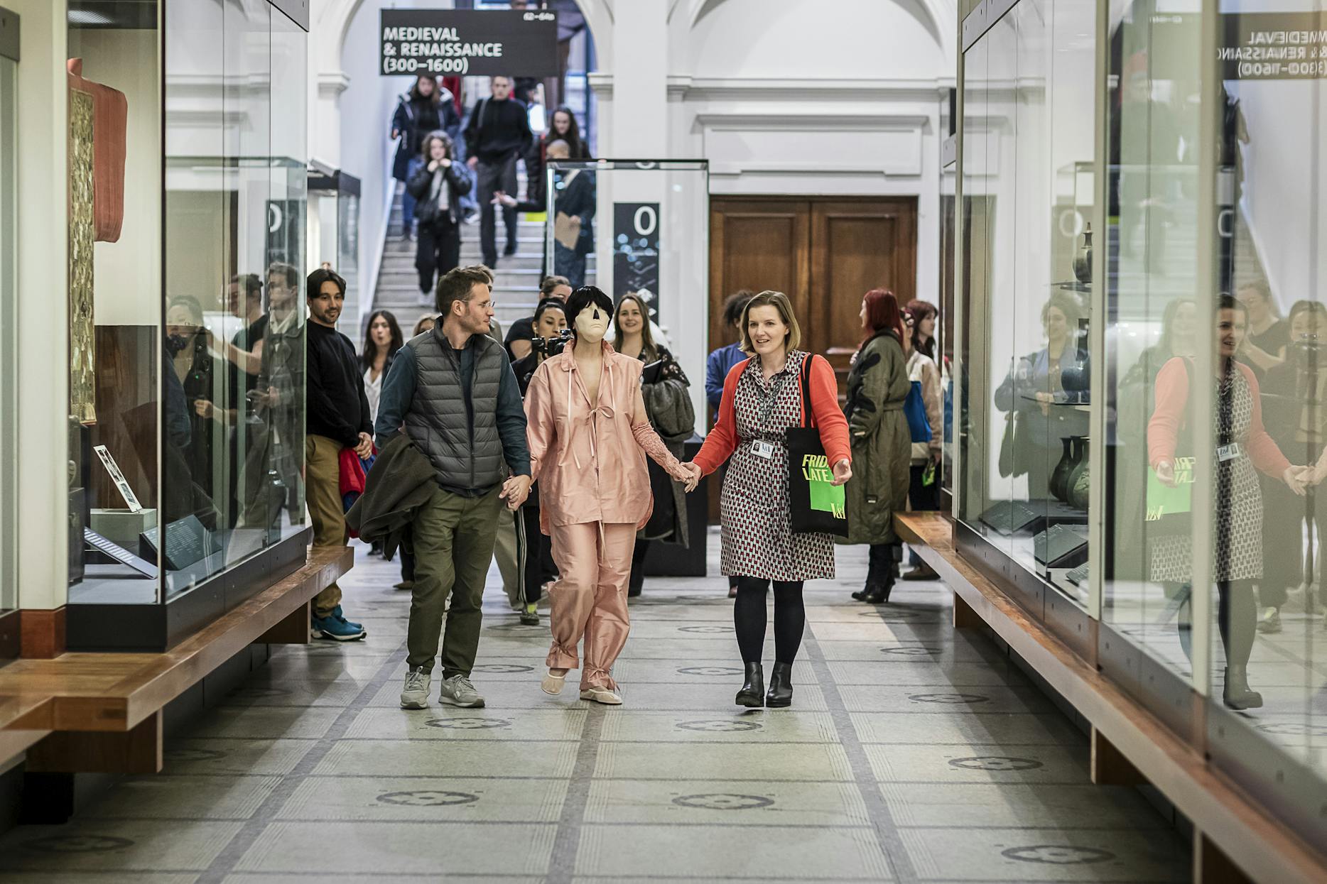 A group of people walk through a museum hallway lined with display cases, with some holding hands and others looking at exhibits; a sign above reads 'Medieval & Renaissance 1300–1600'.