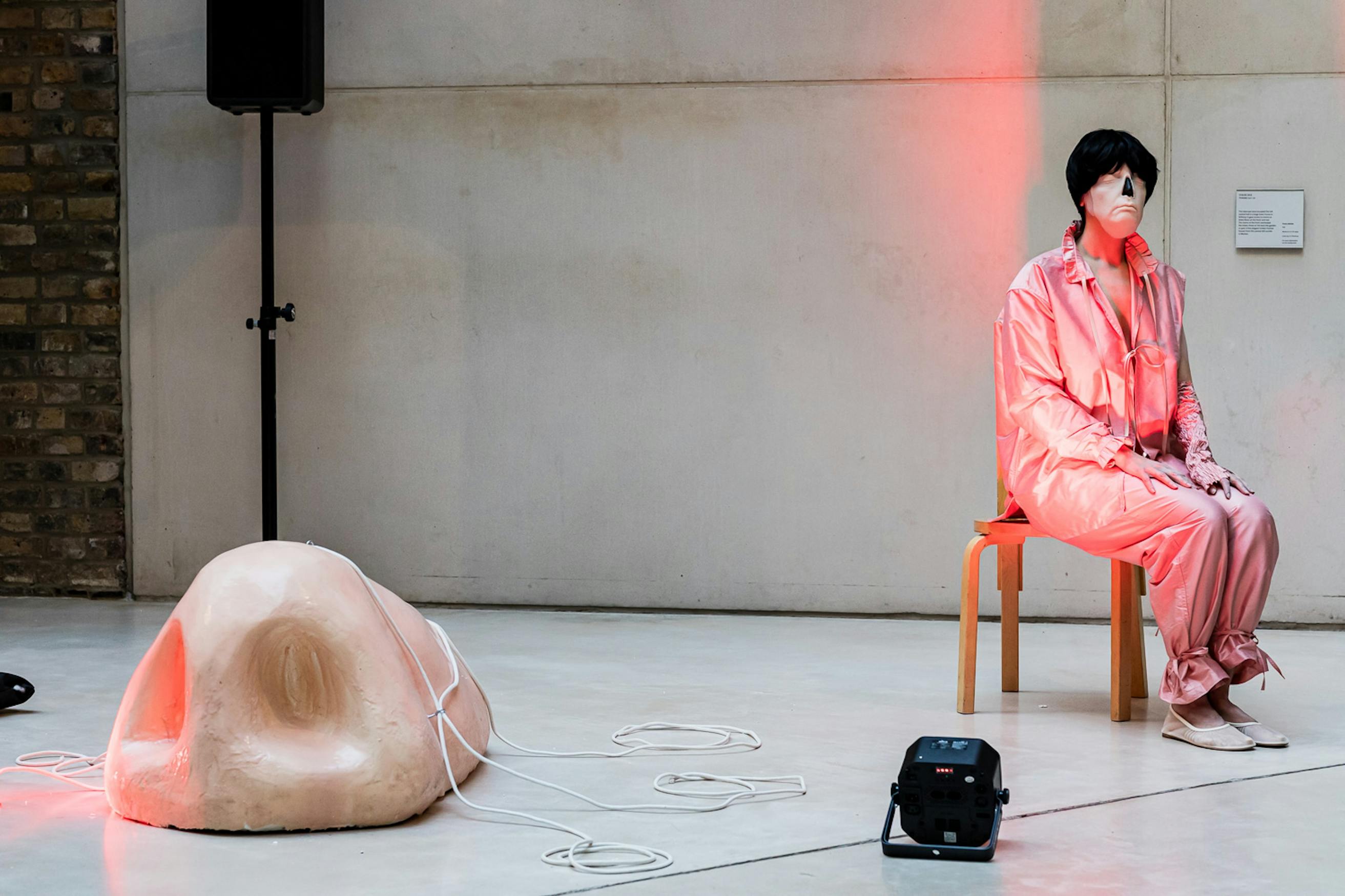 Shirin Fathi in a pink outfit and black wig sits on a wooden chair with a large fake nose on the floor nearby, connected by white cables. The scene is lit with pinkish-red light in a minimalist, industrial space.