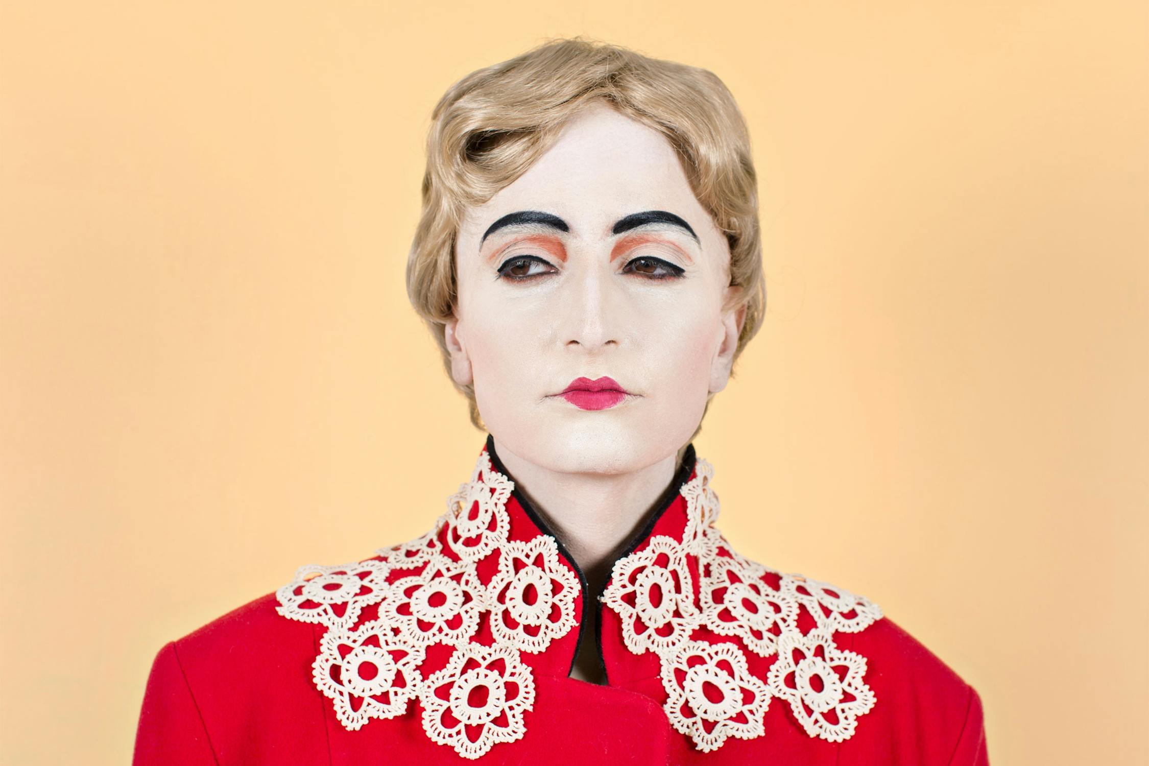 Artist Shirin Fathi with pale makeup, dark arched eyebrows, and red lipstick wears a blonde wig and a red coat with an ornate white lace collar, set against a plain peach background.