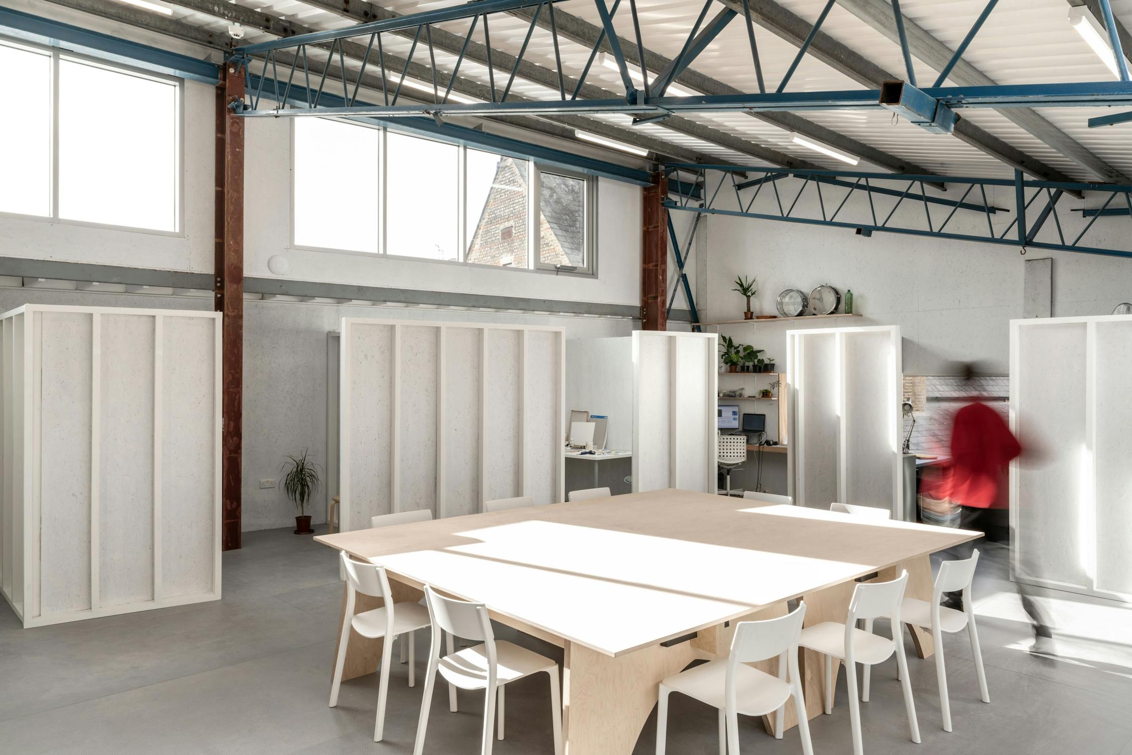 Studio Voltaire's modern, minimalist communal artist studio with white walls, exposed blue steel beams, light wood tables, and white chairs. Partitioned workspaces and plants decorate the room. A person in red walks past, creating a motion blur.