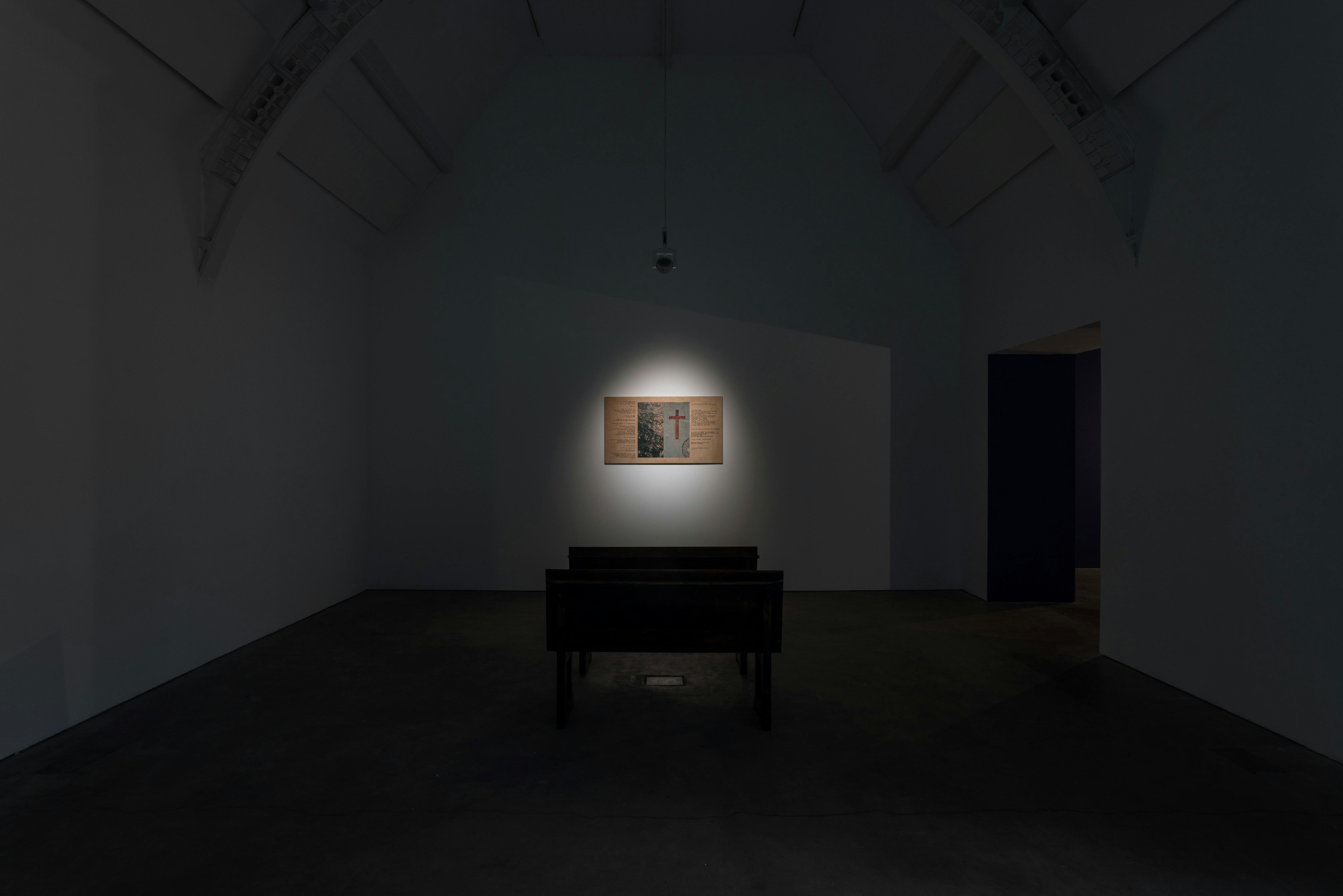 A dimly lit room with pew-like benches facing a single framed artwork on the far wall, illuminated by a spotlight. The space has high, white vaulted ceilings and dark concrete floors.