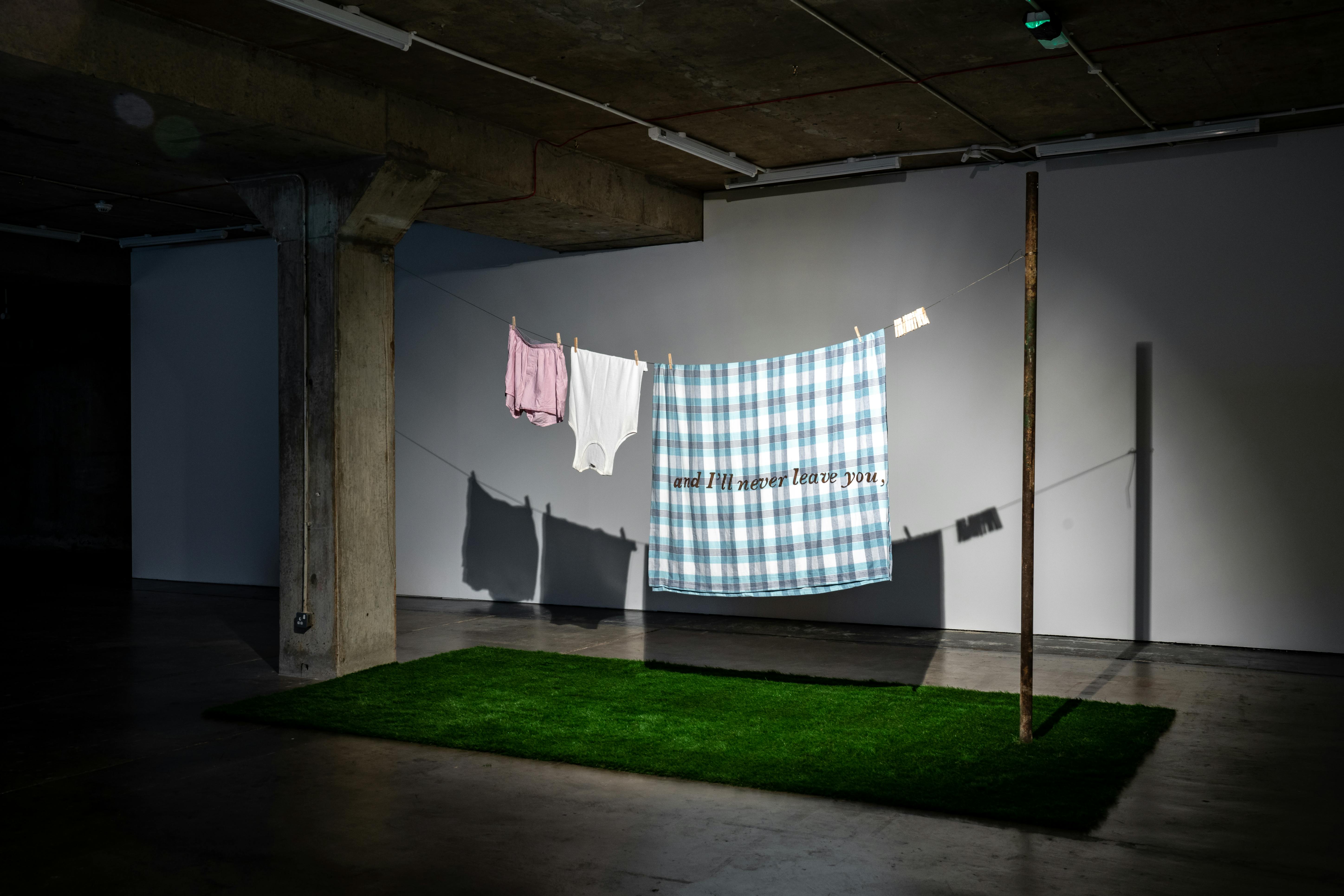Two ropes are strung between a concrete pillar and a pole, holding a pink shirt, white shorts, and a blue plaid blanket with text, above a patch of artificial grass in a dimly lit room with concrete walls and ceiling.