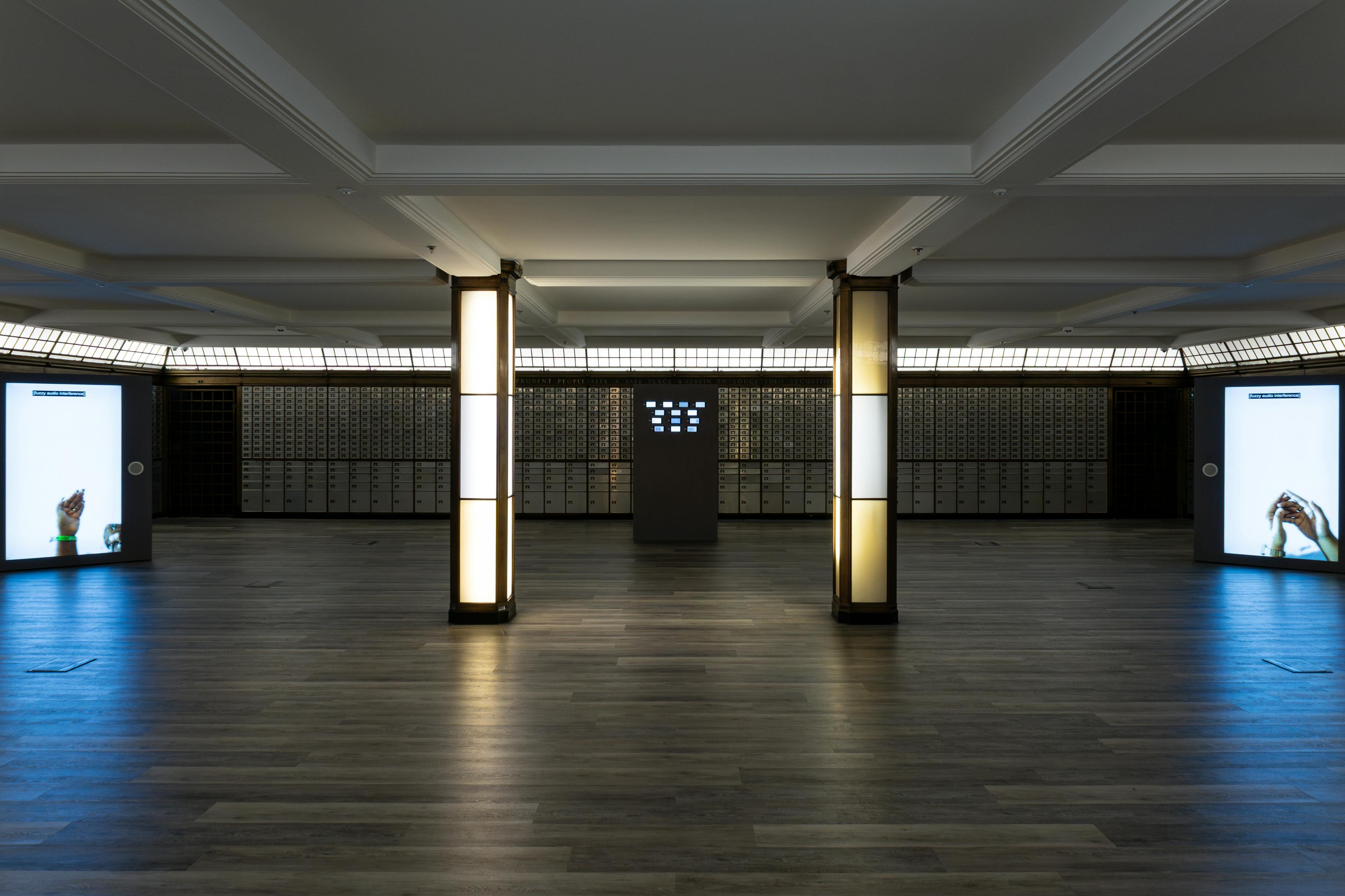 A spacious, modern room with wood flooring, two illuminated pillars, wall-mounted screens displaying hands on each side, and a central kiosk in front of rows of safety deposit boxes.