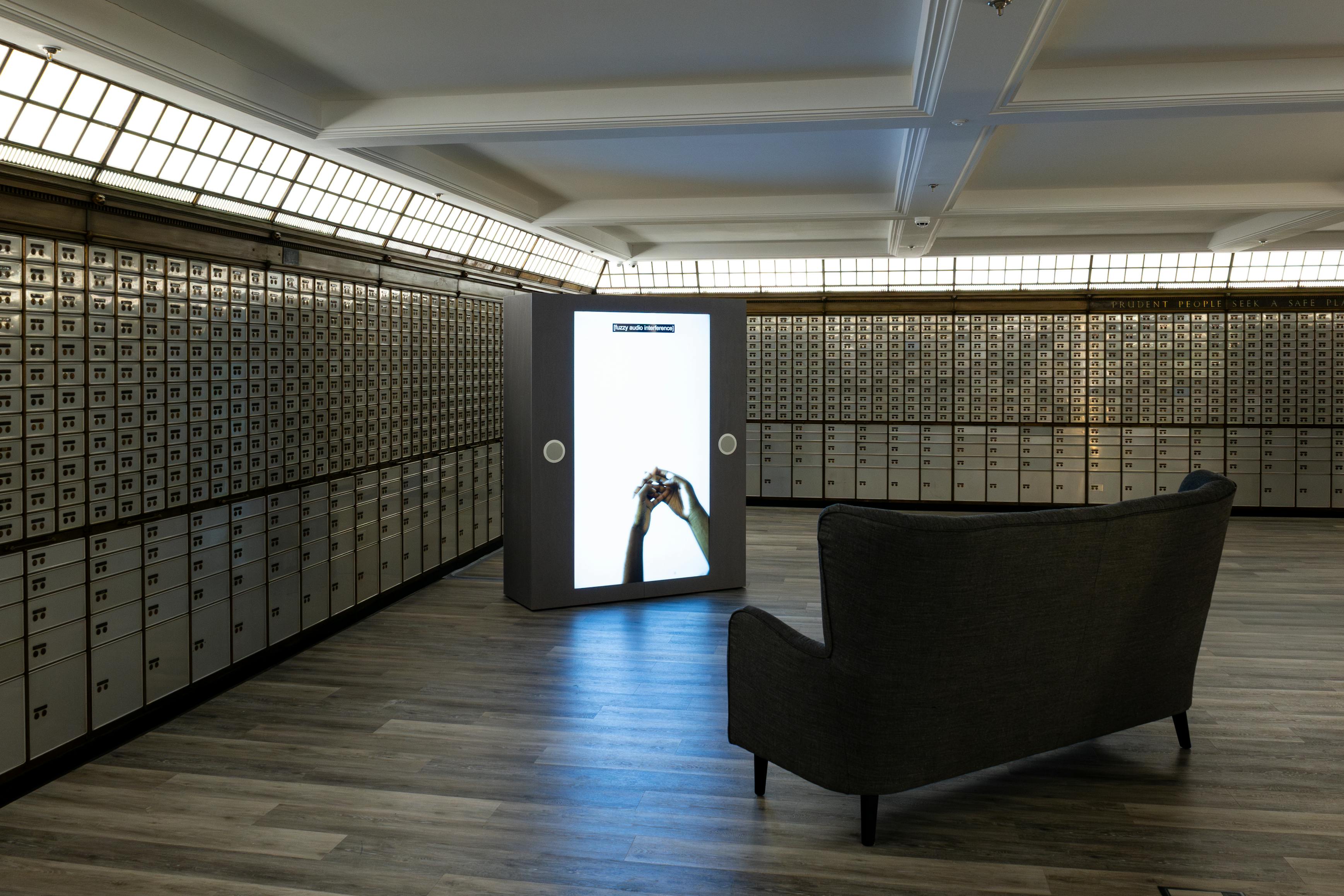 A modern room with rows of metal safe deposit boxes, a dark sofa facing a large, illuminated vertical screen displaying hands holding an object, and wooden flooring throughout the space.