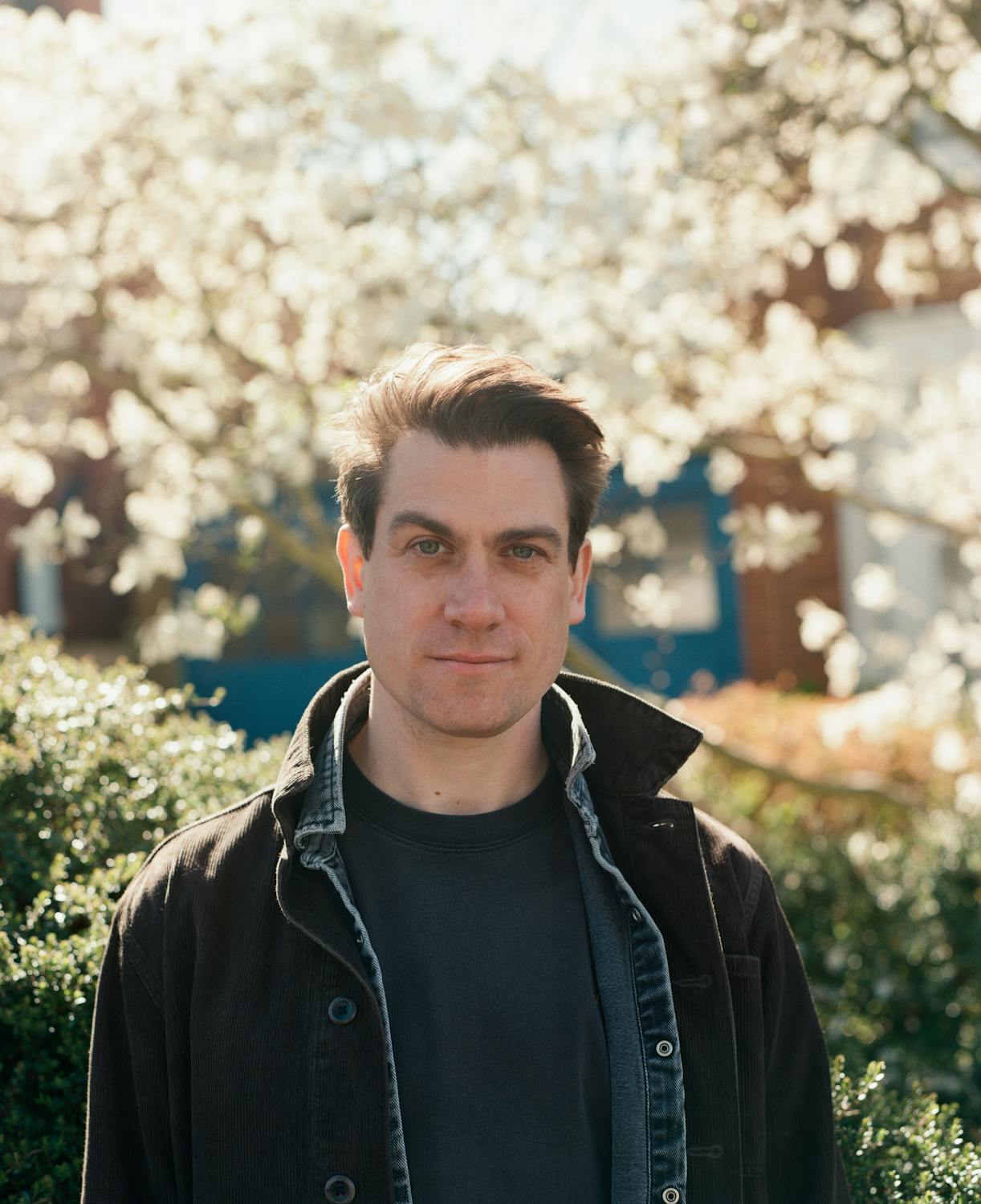 A portrait of Jamie Sutcliffe. A man with short brown hair stands outside in front of blooming trees and bushes, wearing a dark jacket over a T-shirt and looking directly at the camera with a neutral expression.