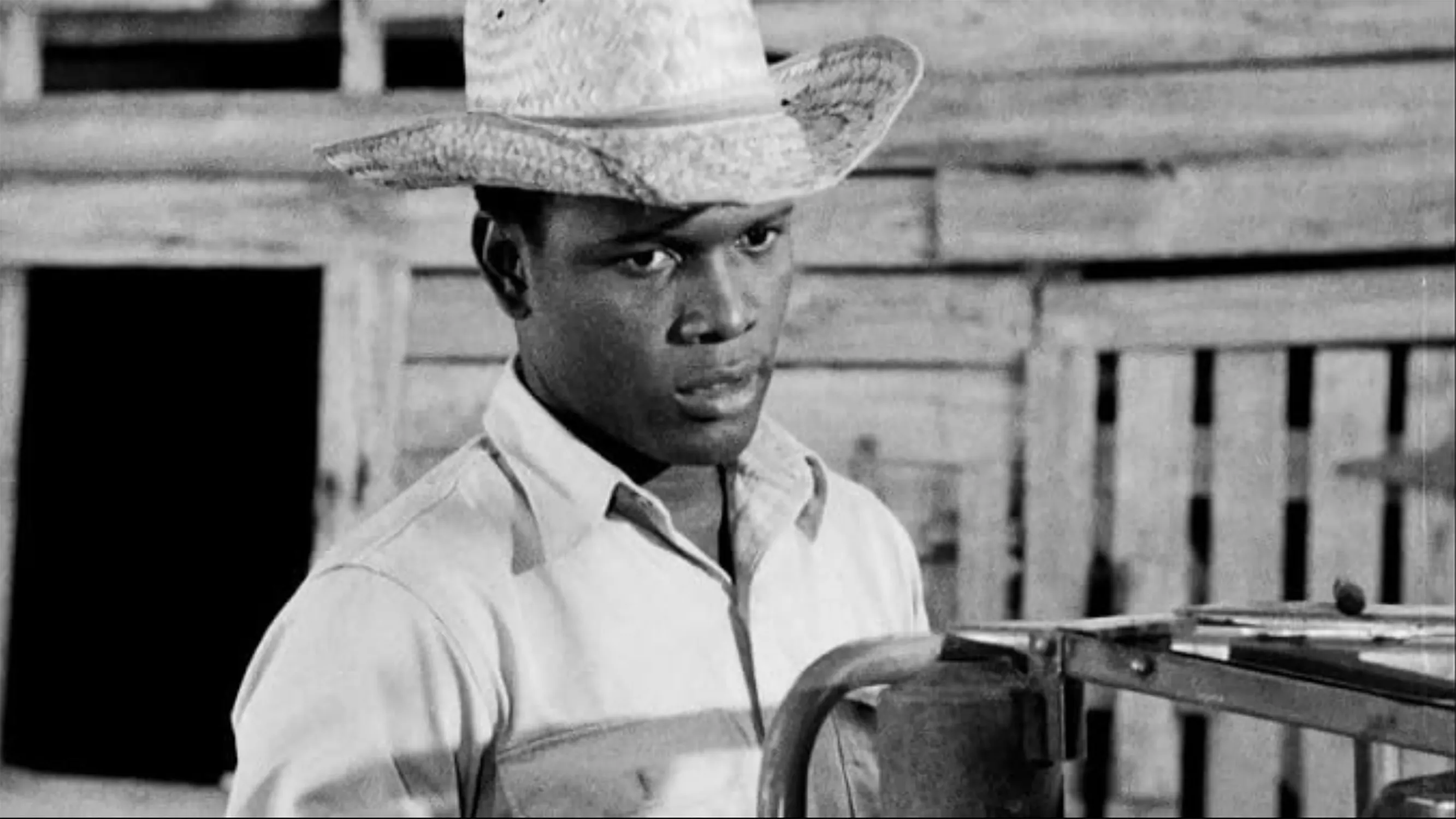 A man in a light-coloured shirt and straw hat stands in front of a rustic wooden building, looking intently to the side. The image is in black and white.
