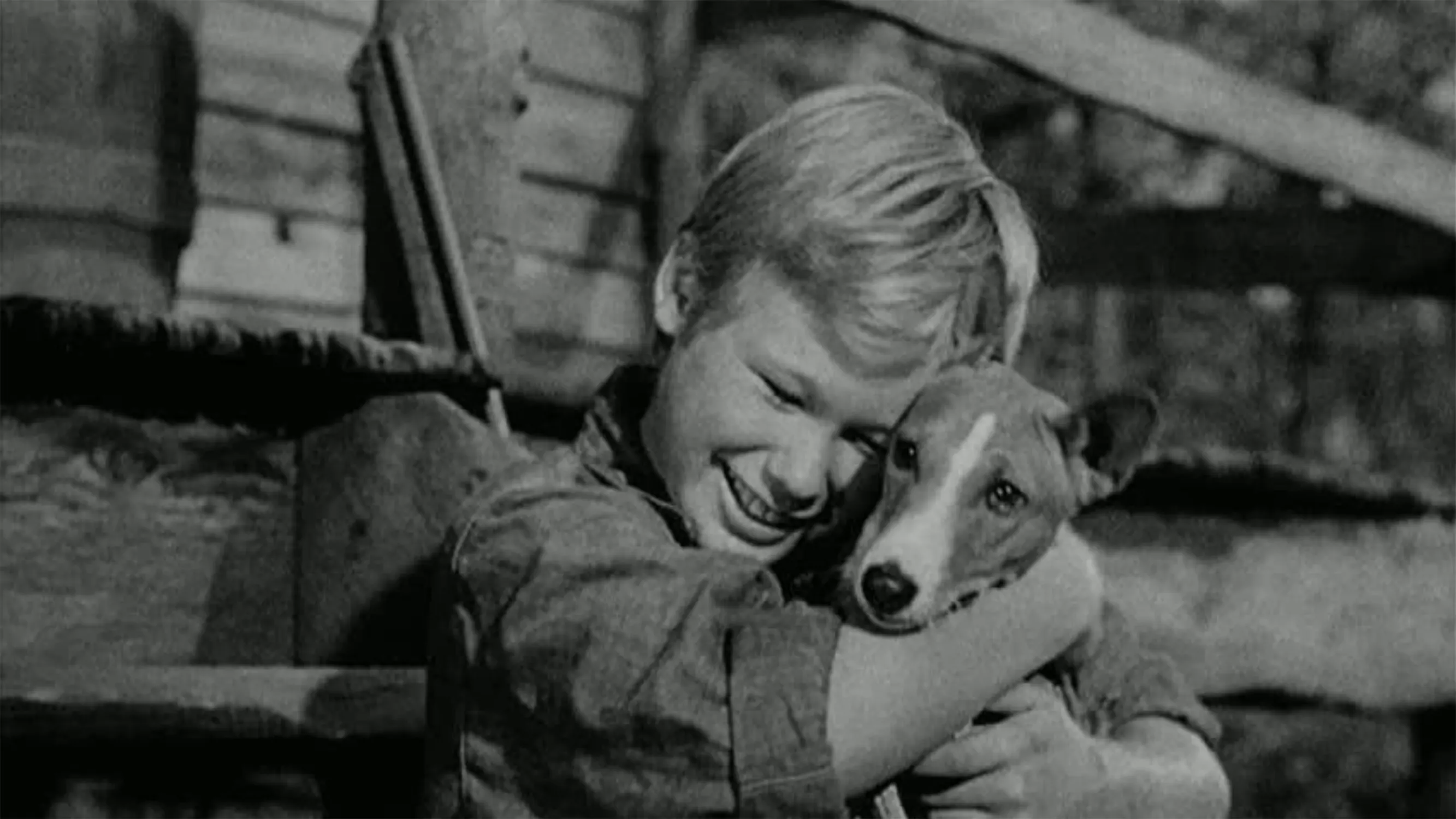 A smiling young boy hugs a dog tightly while sitting outdoors in front of a wooden fence. The image is in black and white, giving it a nostalgic, classic feel.