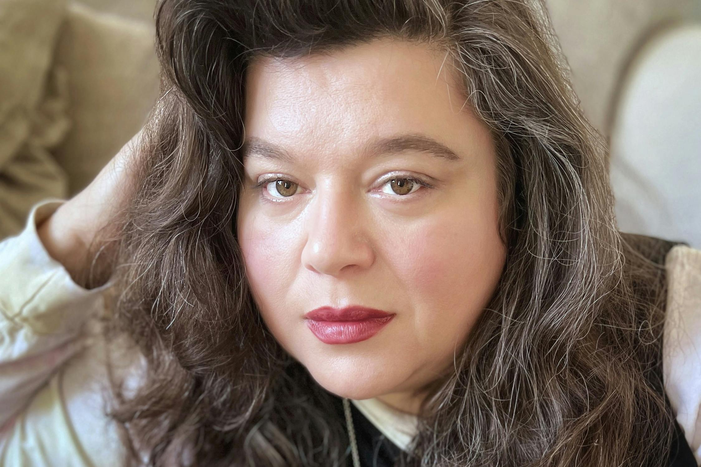 A portrait of Tai Shani: a person with long wavy brown hair and fair skin gazes into the camera with a neutral expression, resting one hand by their head while sitting indoors in soft natural light.