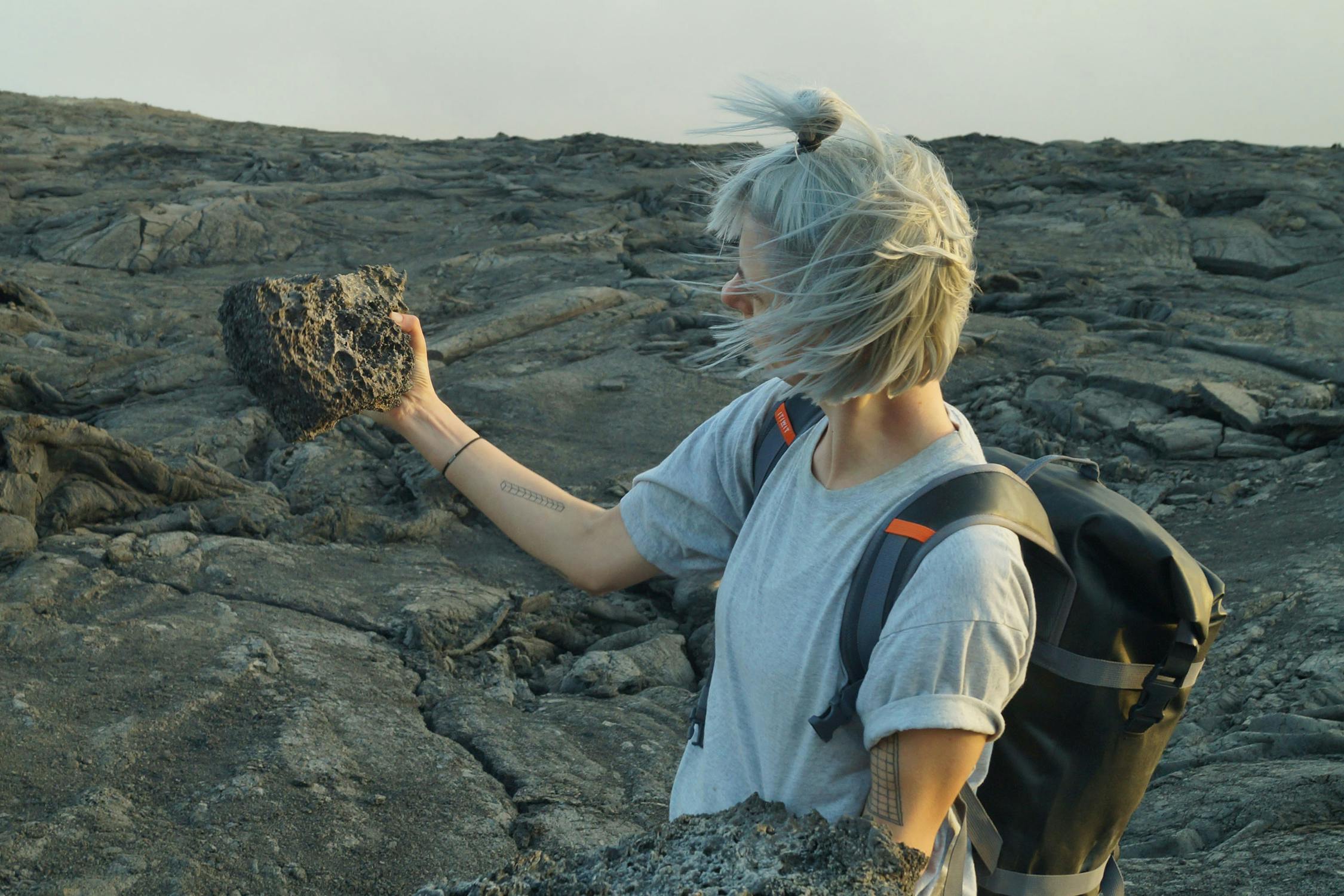 A portrait of Rachel Pimm: a person with short, silver hair and a backpack holds up a large volcanic rock while standing on rough, rocky terrain. Their hair is blown by the wind, and they are wearing a grey t-shirt.