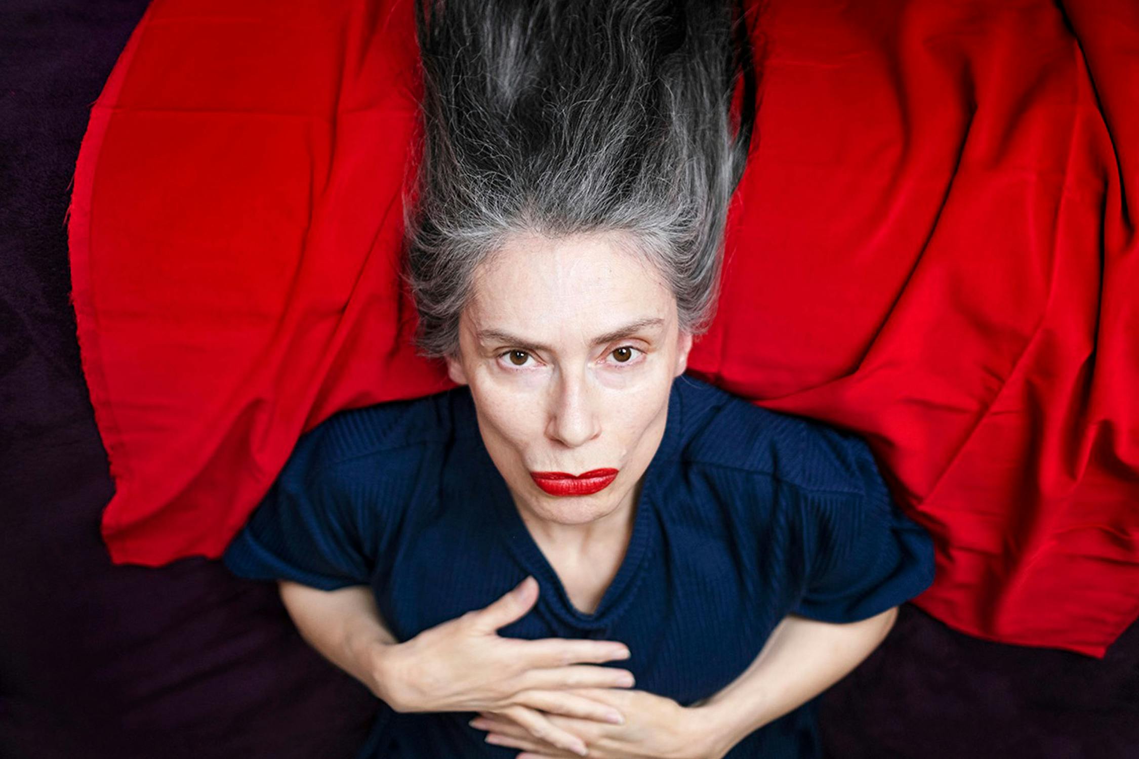 A portrait of Penny Goring, with long grey hair lying on a red cloth, wearing a dark blue top, looking up at the camera with hands resting on their chest and lips painted red.