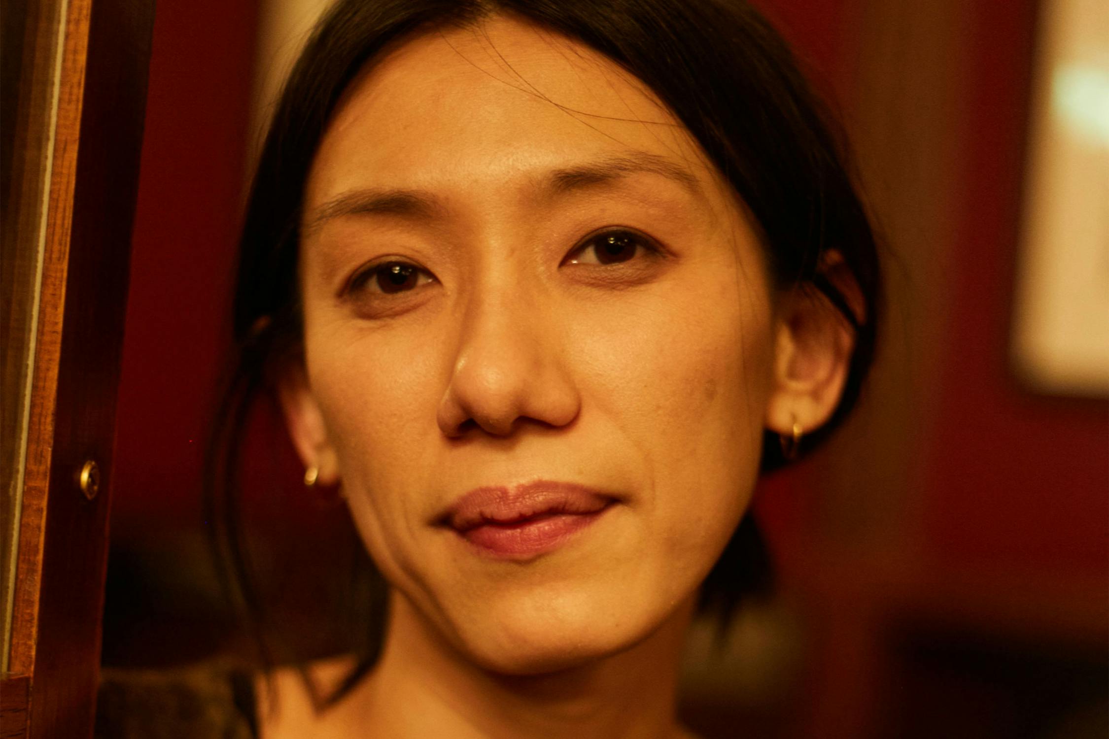 A portrait of Aurelia Guo, with straight dark hair, small hoop earrings, and a neutral expression, looks at the camera in warm, soft indoor lighting with a blurred red and brown background.