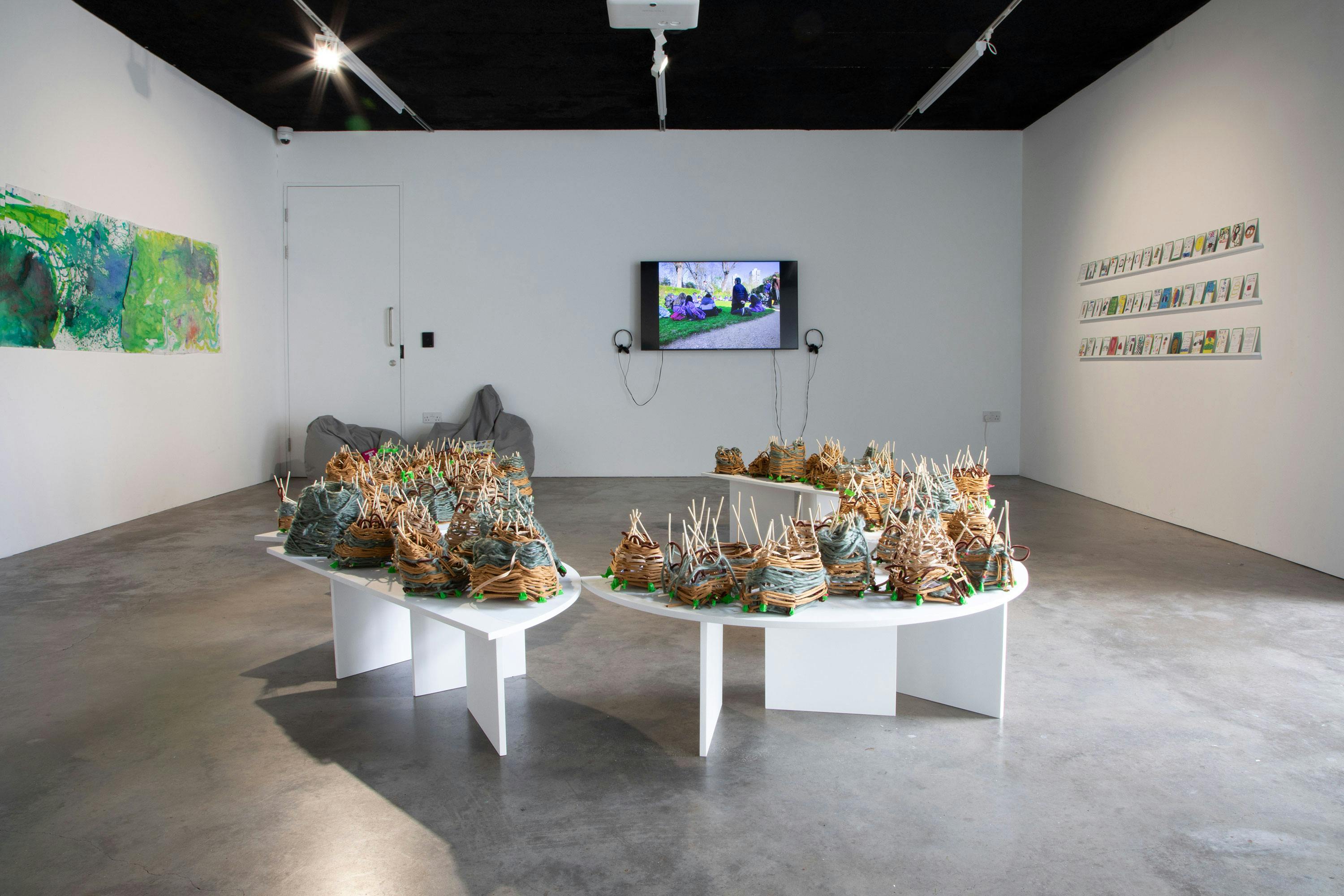 An art gallery with white walls displays colourful woven sculptures on white tables, a wall-mounted screen with headphones, a green abstract painting, and a collection of small hand-drawn playing cards on the right wall.