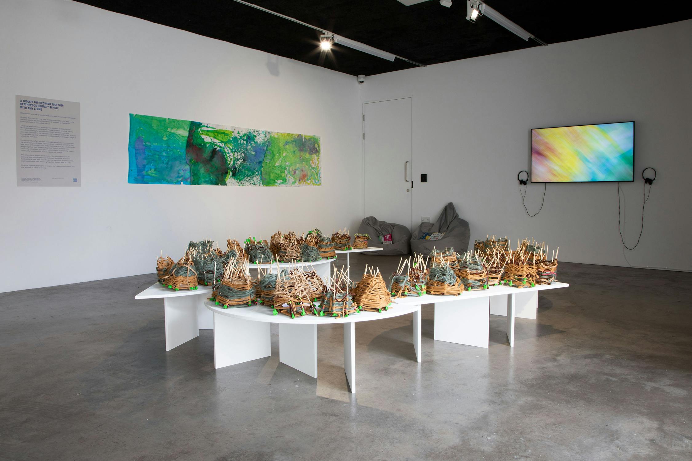 A modern gallery displays woven basket sculptures on white tables, a colourful abstract painting on the wall, a digital screen with headphones, and two grey bean bags in a minimalist space with concrete floors.