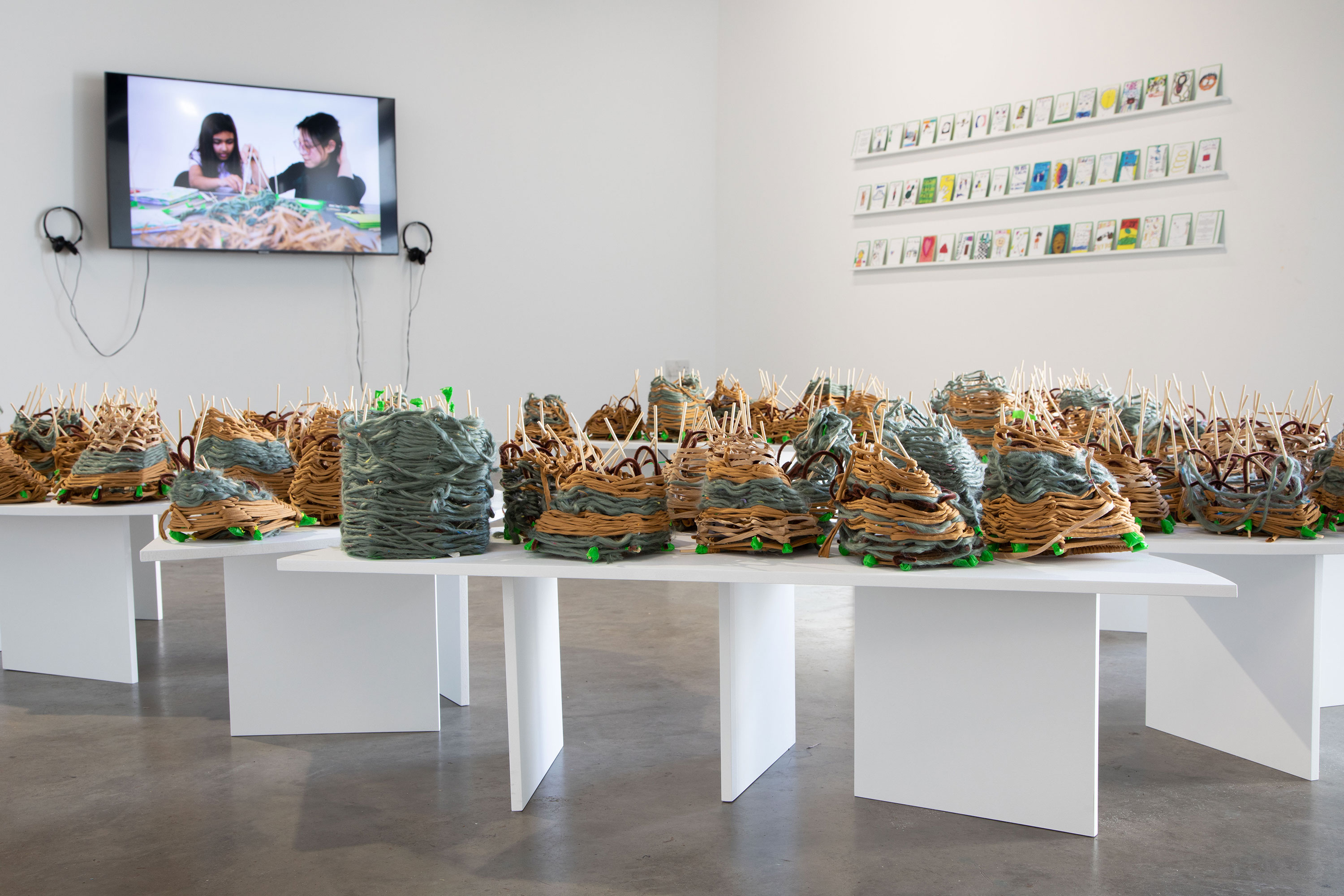 A contemporary art gallery displays clustered abstract sculptures made of layered materials on white tables. On the wall, a video of two people working is shown beside a grid of framed pieces. The space is minimal and bright.