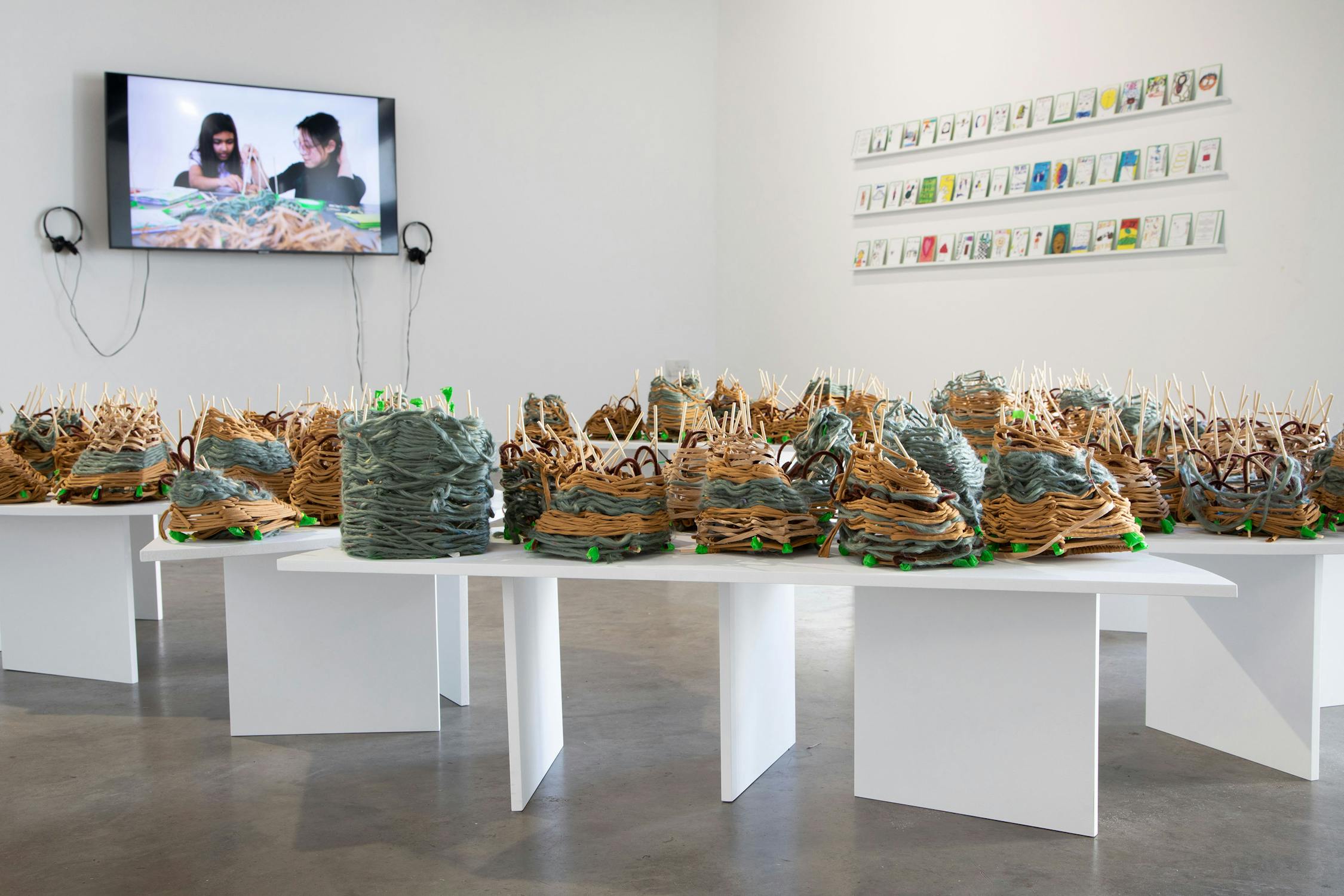 A contemporary art gallery displays clustered abstract sculptures made of layered materials on white tables. On the wall, a video of two people working is shown beside a grid of framed pieces. The space is minimal and bright.