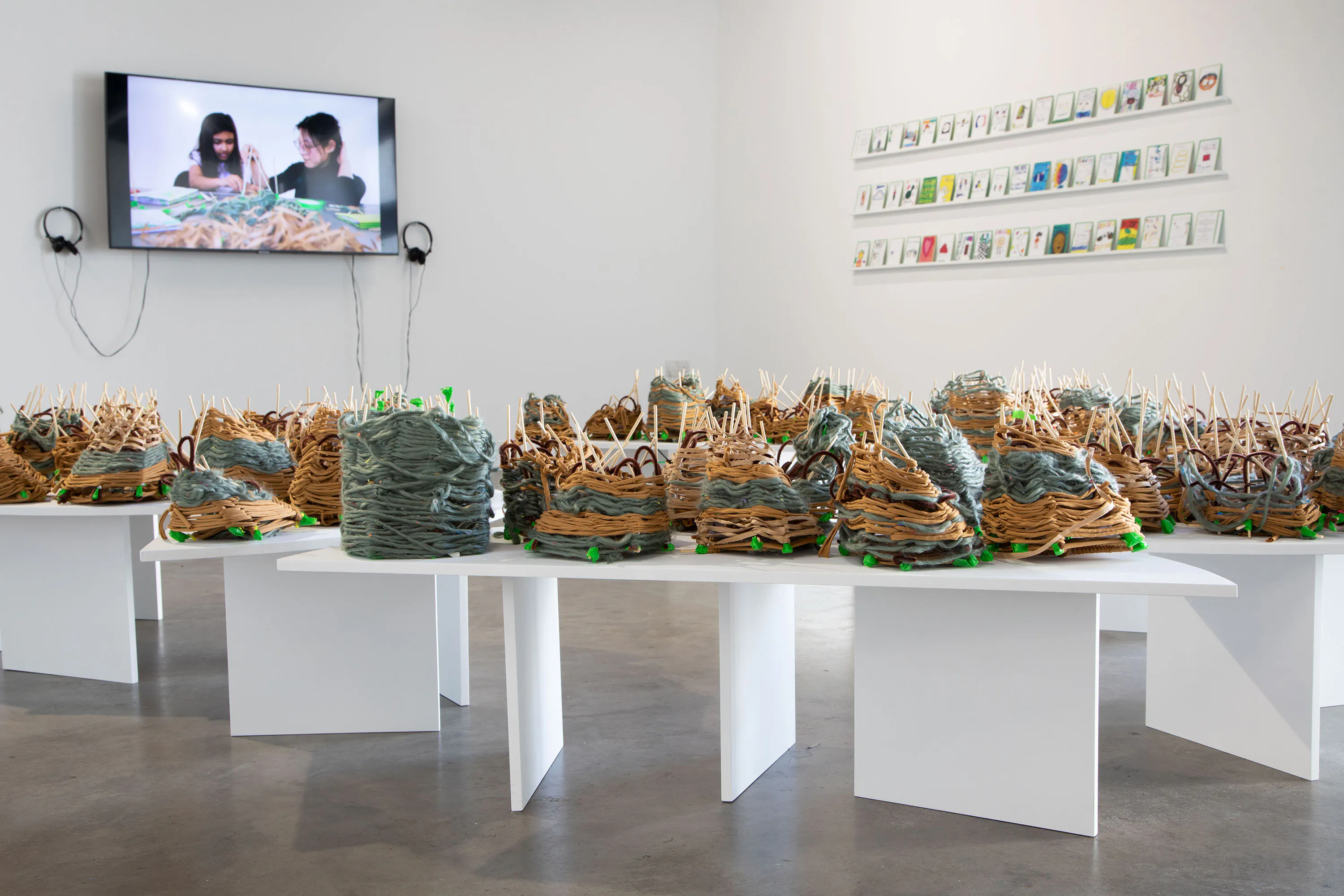 A contemporary art gallery displays clustered abstract sculptures made of layered materials on white tables. On the wall, a video of two people working is shown beside a grid of framed pieces. The space is minimal and bright.