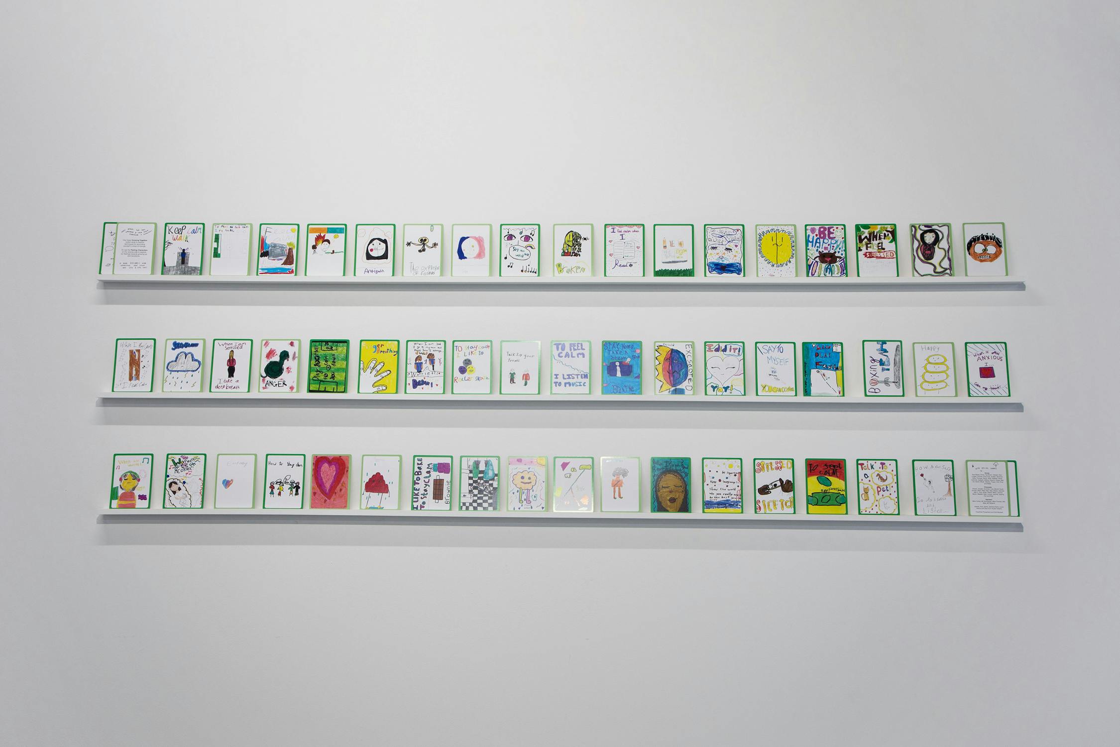 A white wall displays three horizontal shelves, each holding a row of small, colourful cards with unique drawings and handwritten notes. 