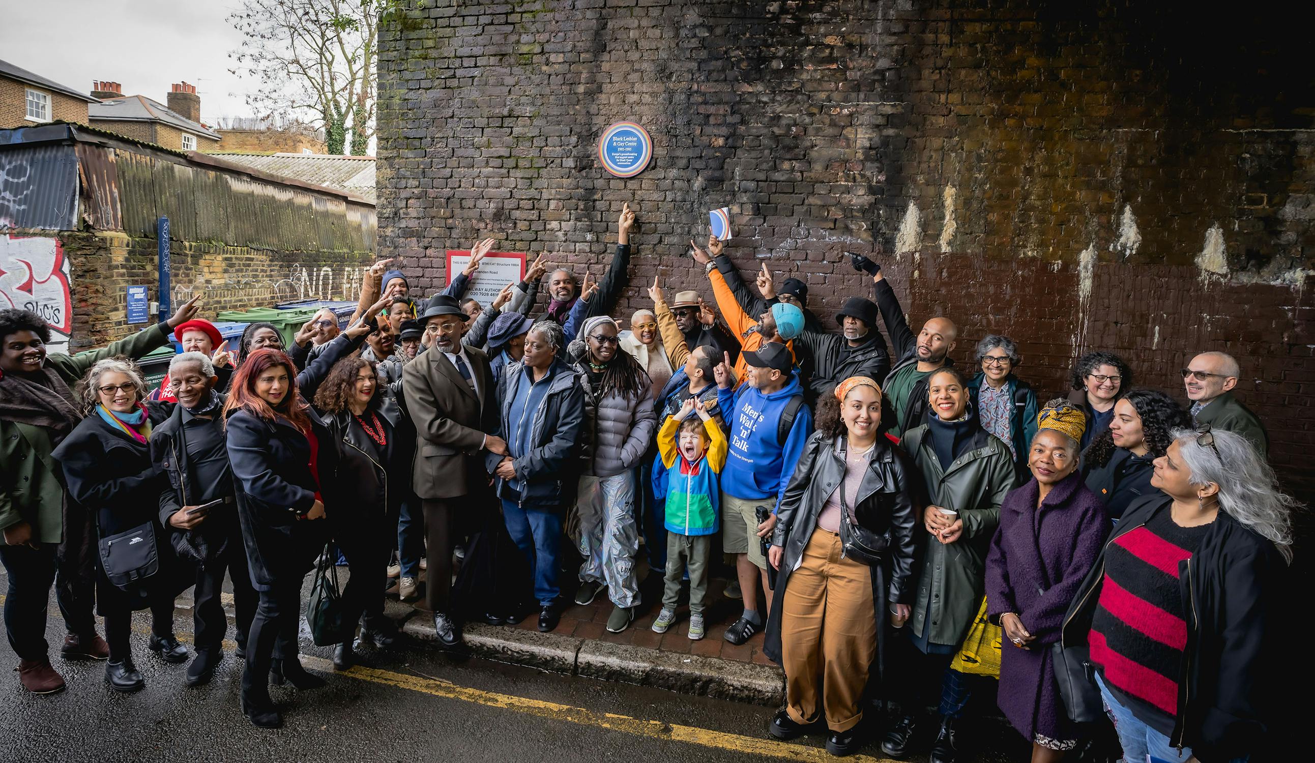 A diverse group of people stand smiling and posing under a brick bridge, some pointing up at a rainbow plaque dedicated to the Black, Lesbian & Gay Centre, on the wall above them. Several people hold up phones or cameras, and the mood appears celebratory.
