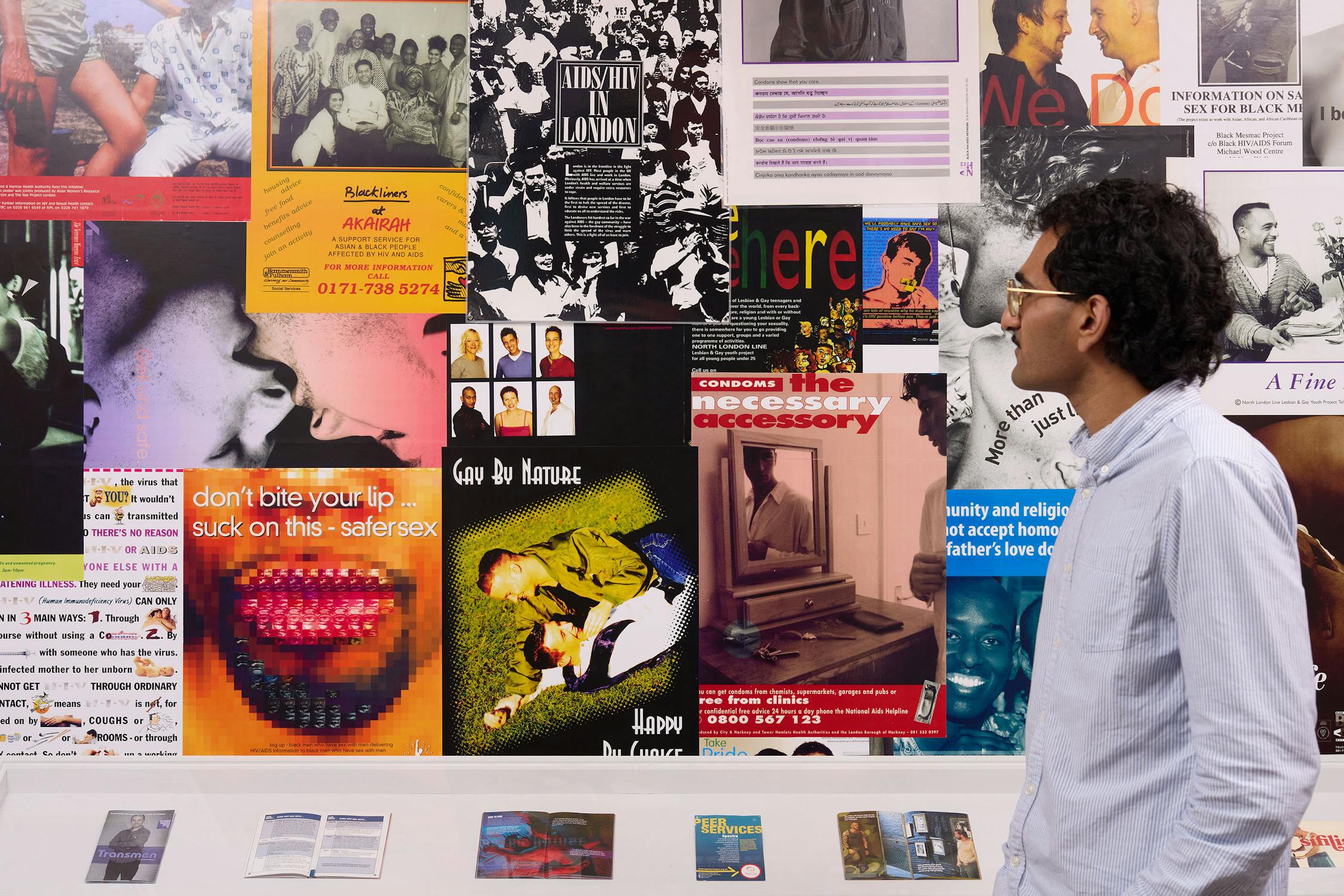 A person in glasses and a light blue shirt looks at a colourful wall covered with various posters, photos, and magazine pages on topics including sexual health and safe sex. Magazines are displayed in a vitrine below.