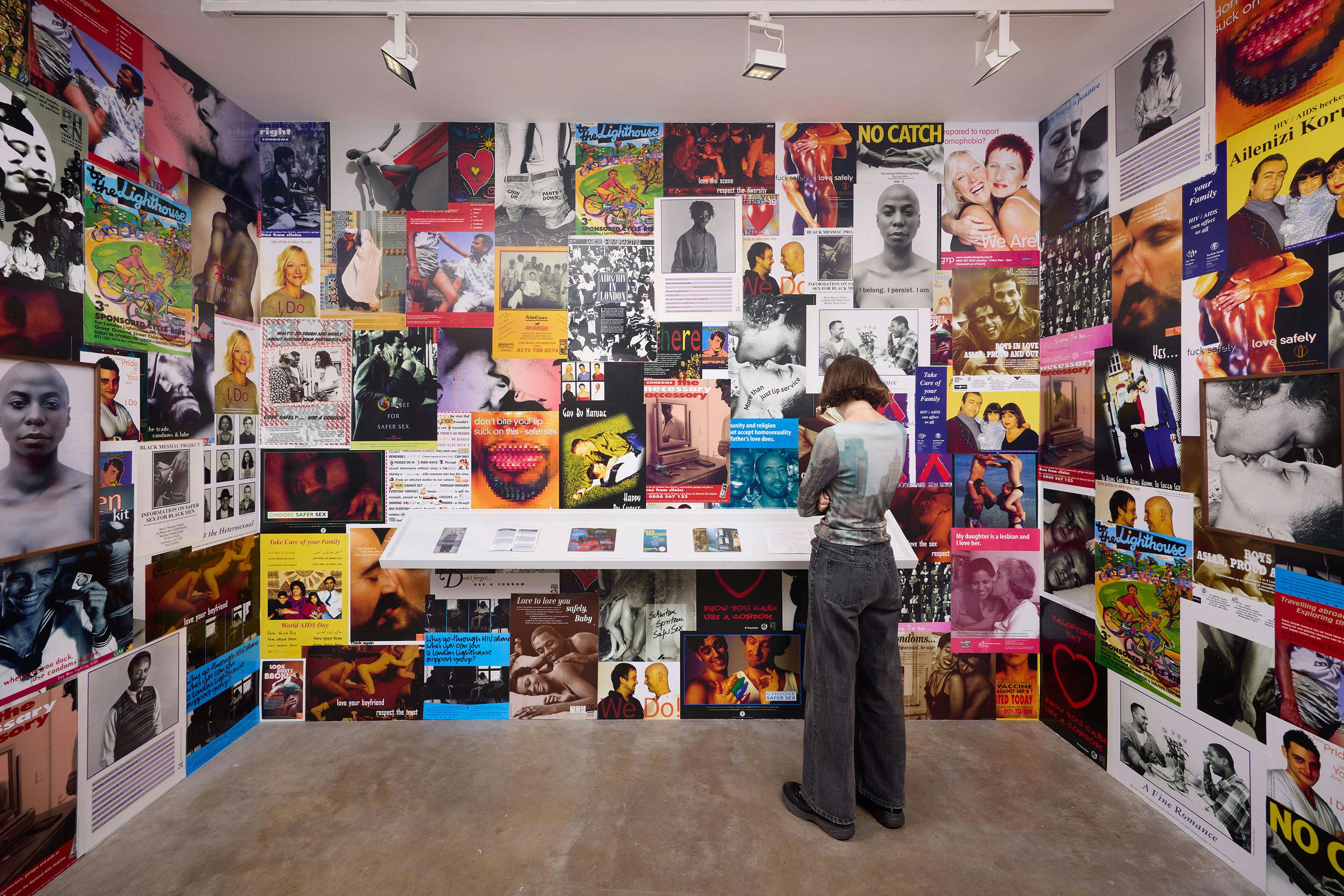 Installation view of It’s A Love Thing at Studio Voltaire. A person stands in front of a colourful wall collage of posters, photos and flyers exploring queer love, identity and sexual health. Presented by Spectra CIC and Studio Voltaire. Photo by Tom Carter.