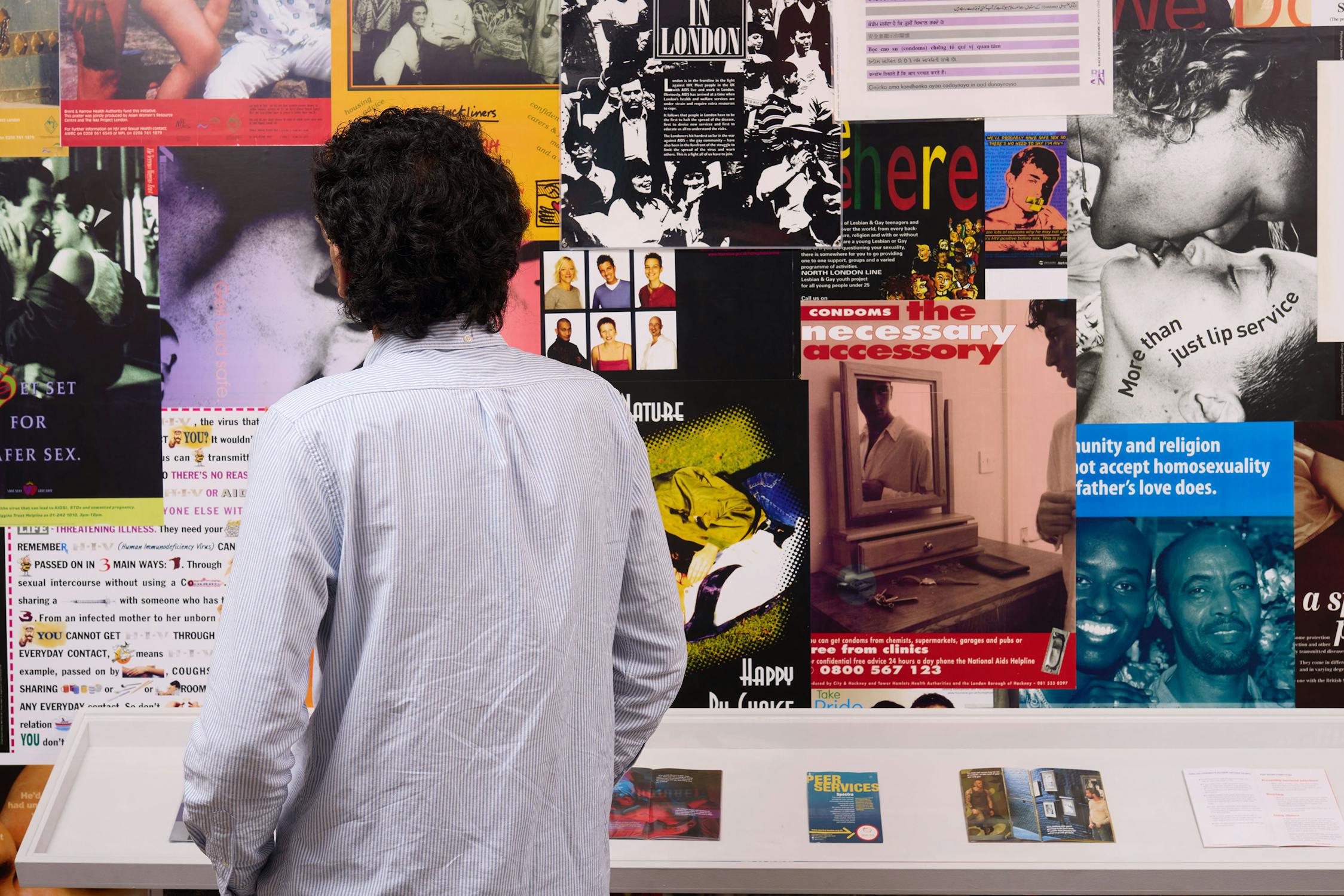 A person wearing a light blue shirt looks at a colourful wall covered with various posters, photos, and magazine pages on topics including sexual health and safe sex. Magazines are displayed in a vitrine below.