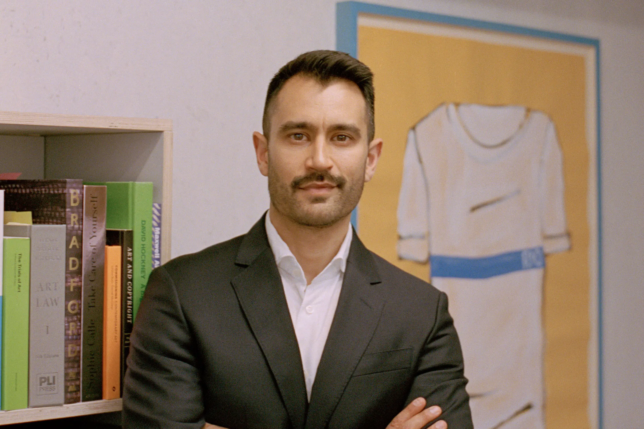 Adam Jomeen, an art lawyer, stands with arms crossed in front of a bookcase filled with books on art and law. He is wearing a dark suit jacket and white shirt, and is positioned next to a framed artwork depicting a stylised item of clothing.