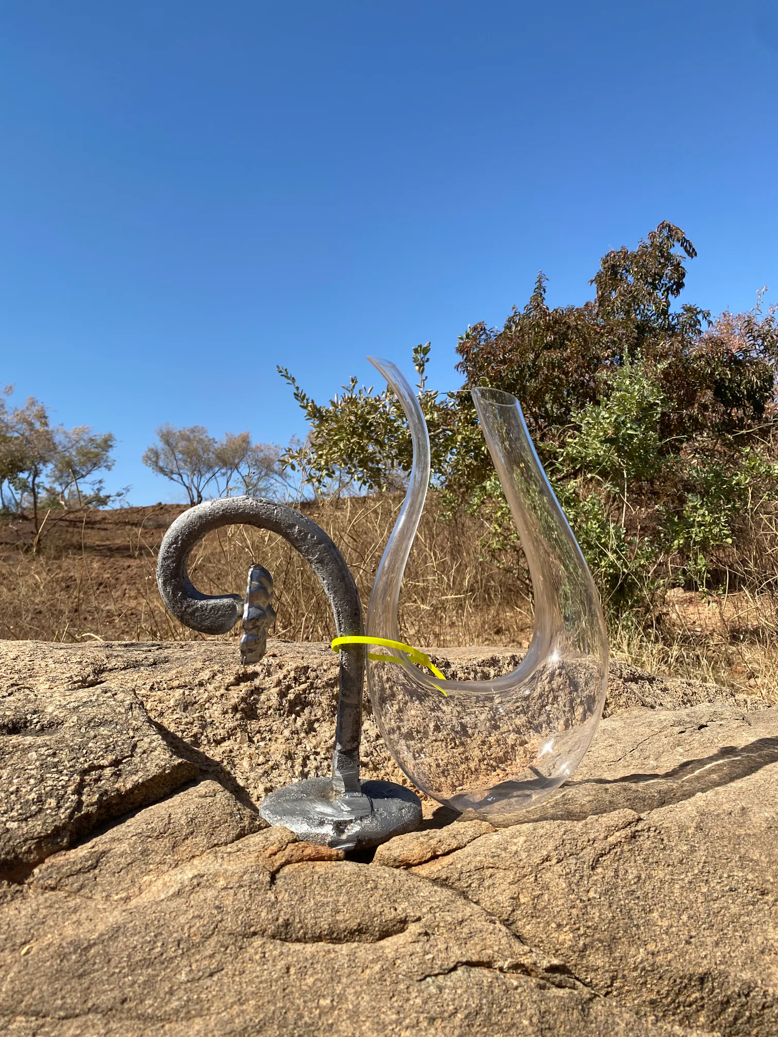 A clear, curved glass vessel sits on a rocky surface outdoors, next to a metal stand with a hook, under a clear blue sky with some trees and shrubs in the background.